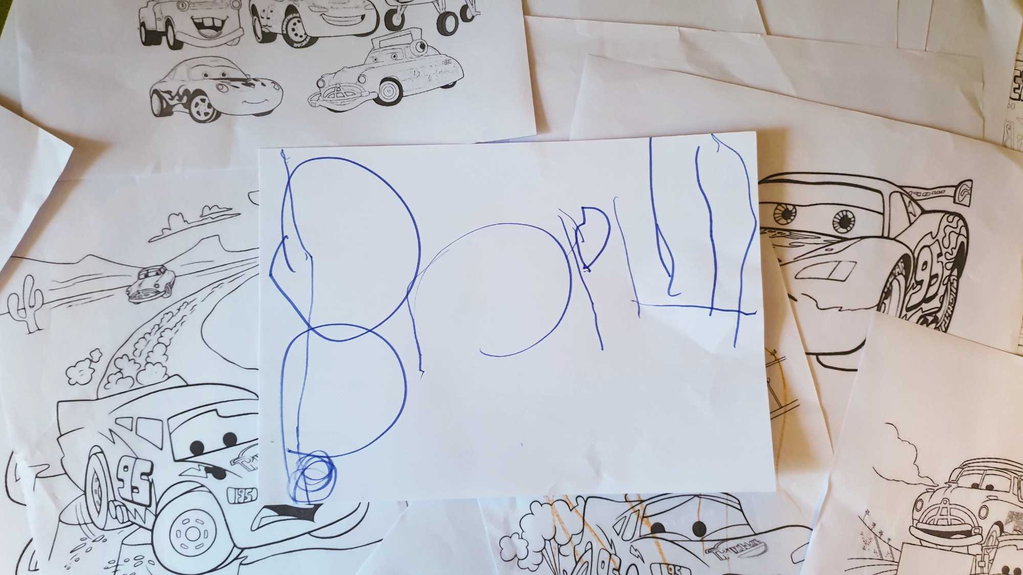 На белой бумаге синим маркером написано слово «Ворл», лежащее на фоне раскрасок с персонажами мультфильма «Тачки».