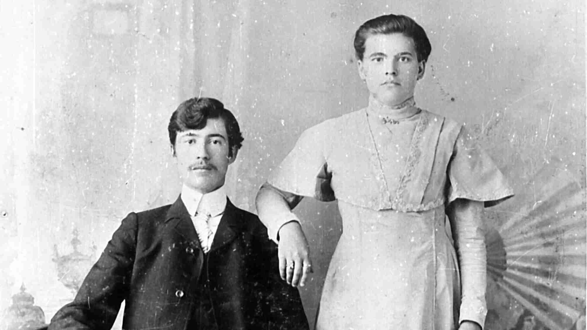 Чёрно-белая фотография: мужчина в тёмном костюме сидит слева, женщина в светлом платье стоит справа, её рука на его плече; фон — студийный с радиальным узором.