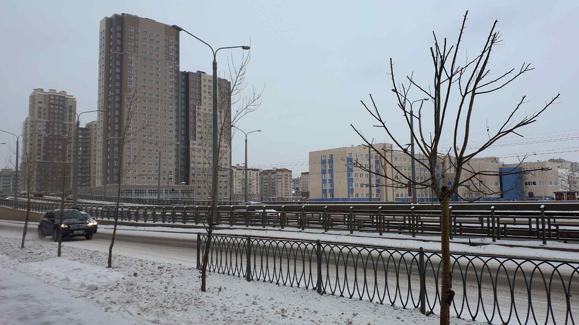 Зимний городской пейзаж: заснеженная дорога с движущимися автомобилями, высокие жилые дома на фоне пасмурного неба, голые деревья и металлические ограждения.