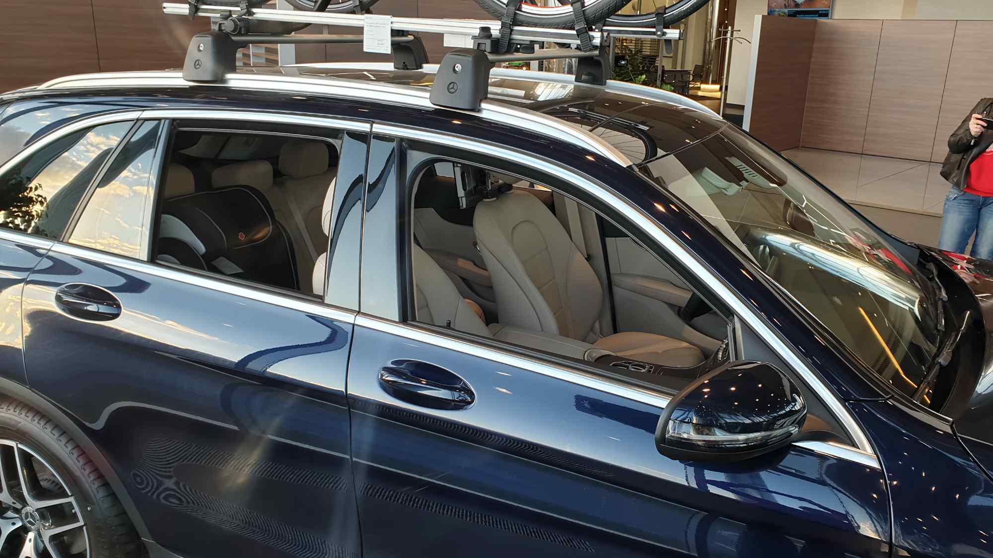 Тёмно-синий универсал Mercedes с багажником на крыше и велосипедом внутри автосалона, видны светлые кожаные сиденья и часть человека справа.