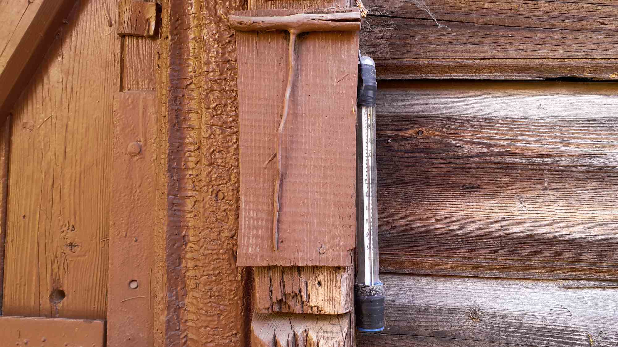 Уличный термометр, закреплённый на деревянной стене с видимой текстурой и трещинами, показывает температуру около 20°C.