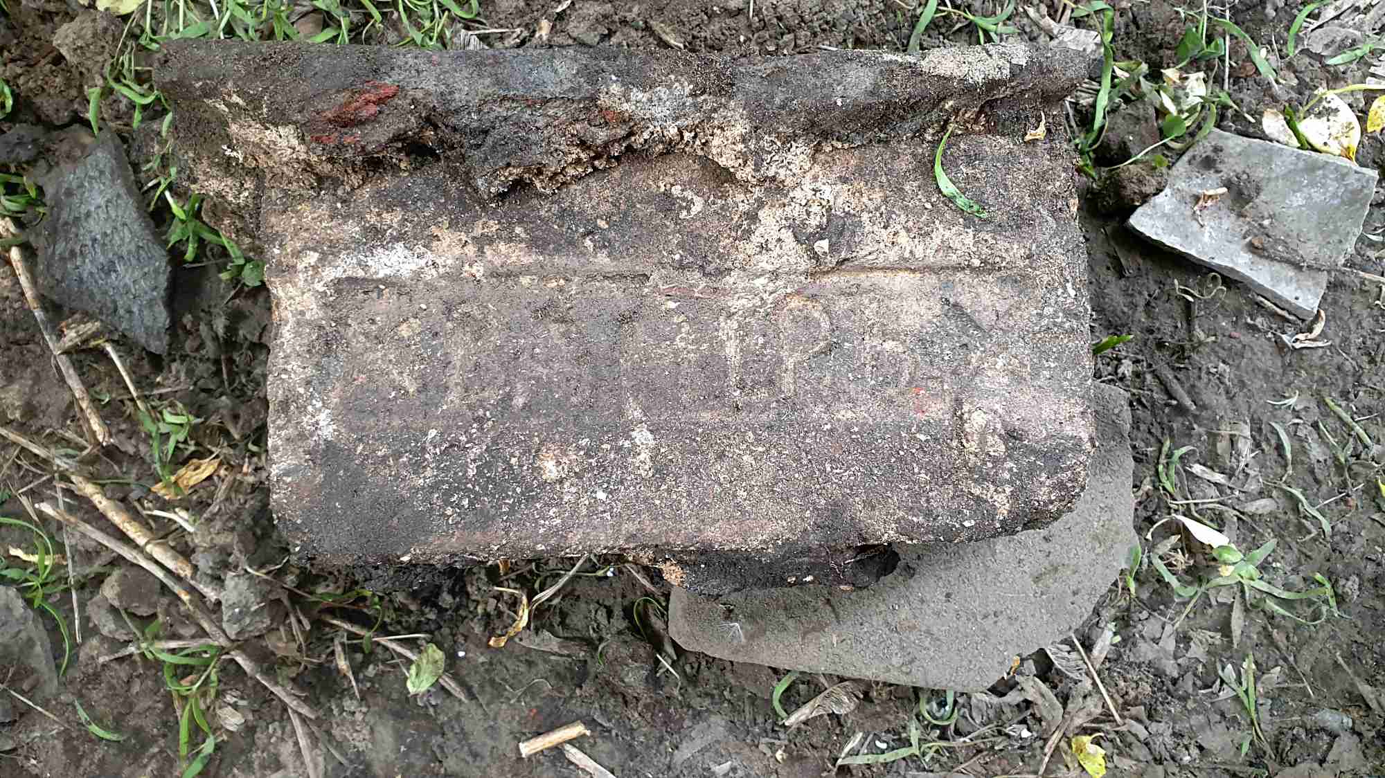 Старый кирпич с надписью «1890» лежит на земле среди травы и мусора.