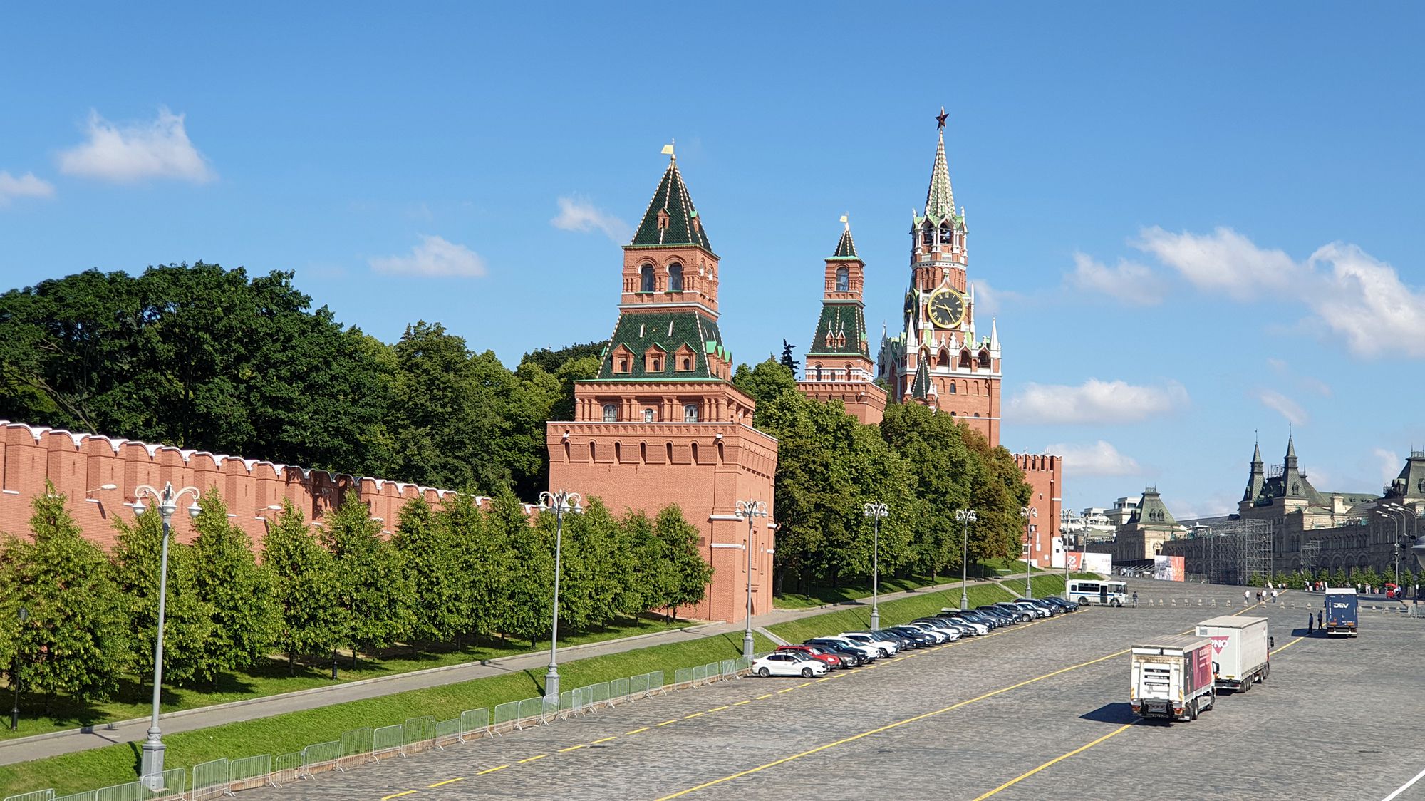 Вид на Московский Кремль: красные стены, башни с зелёными крышами и звездой, Спасская башня с часами, парковка и грузовики на площади под голубым небом.