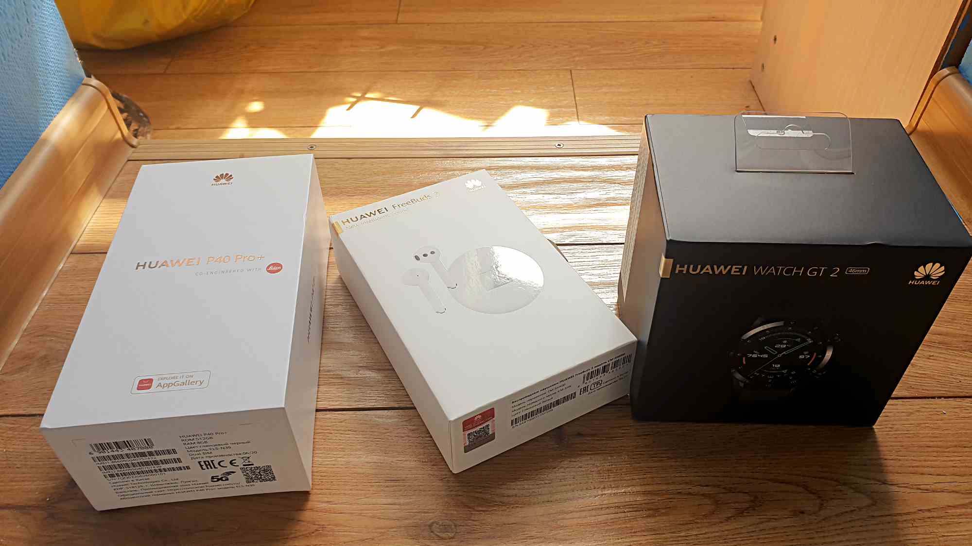 Три коробки Huawei: P40 Pro+, FreeBuds 3 и Watch GT 2, расположенные на деревянном полу под солнечным светом.
