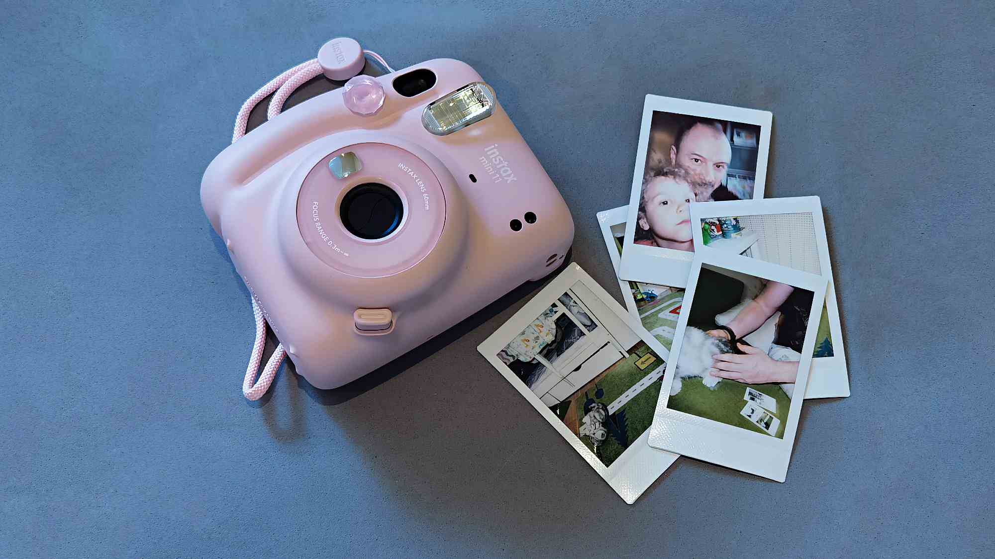 Розовая мгновенная камера Fujifilm Instax Mini 11 с розовым шнурком лежит на сером фоне рядом с несколькими отпечатанными фотографиями, включая снимки людей и животных.