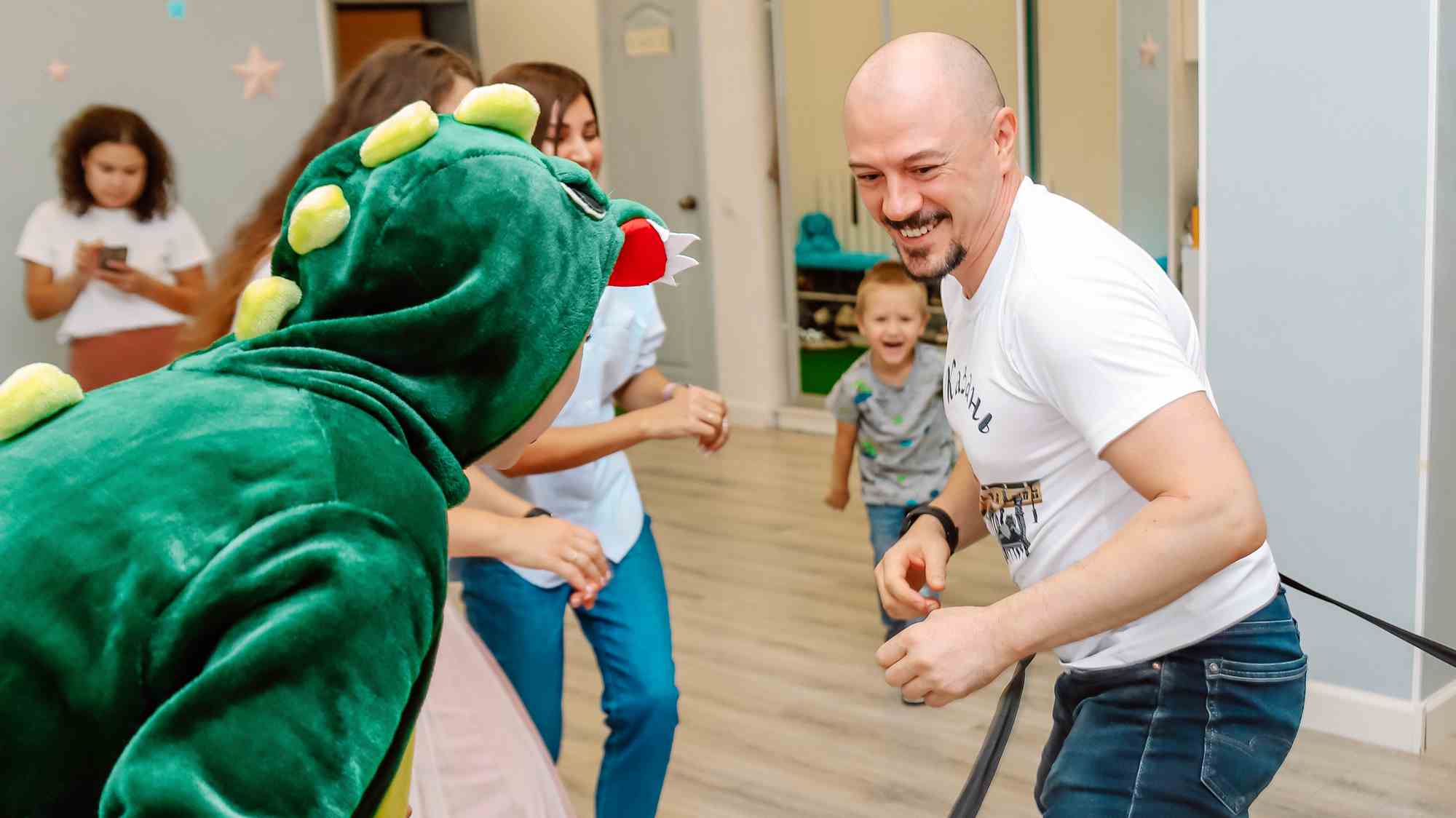 Мужчина в белой футболке с надписью «Казань» и ребёнок в зелёном костюме динозавра играют в помещении, улыбаясь друг другу; на заднем плане — другие дети и взрослые.