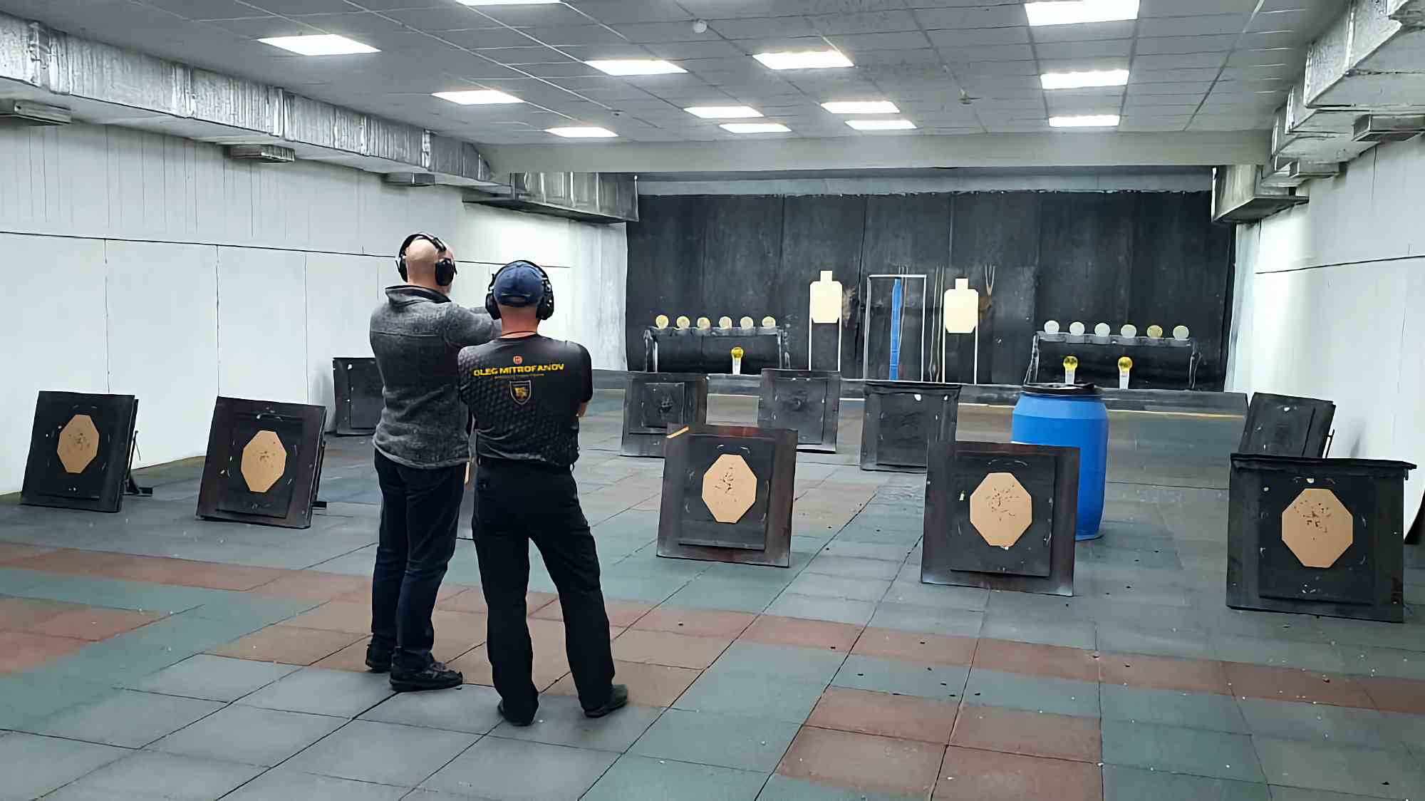 Два человека в наушниках на стрельбище: один в серой кофте стреляет, другой в футболке «OLEG MITROFANOV» наблюдает; видны мишени, барьеры и синий бочонок.