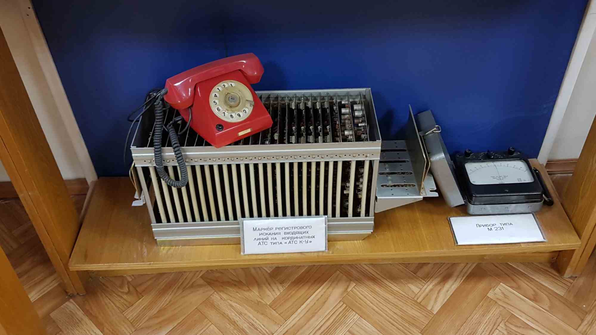Ретро-телефон на металлическом устройстве с надписью «Маркёр регистрового искания входящих линий на координатных АТС типа «АТС К-У» и прибор типа М-231 на деревянной полке.