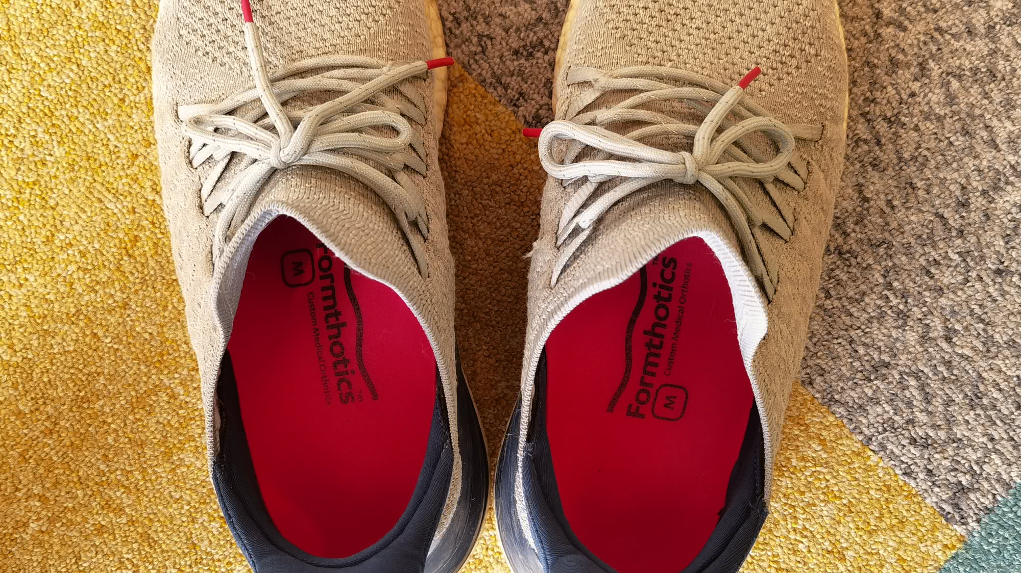 Бежевые кроссовки с красными стельками Formthotics Custom Medical Orthotics, вид сверху на разноцветном ковре.