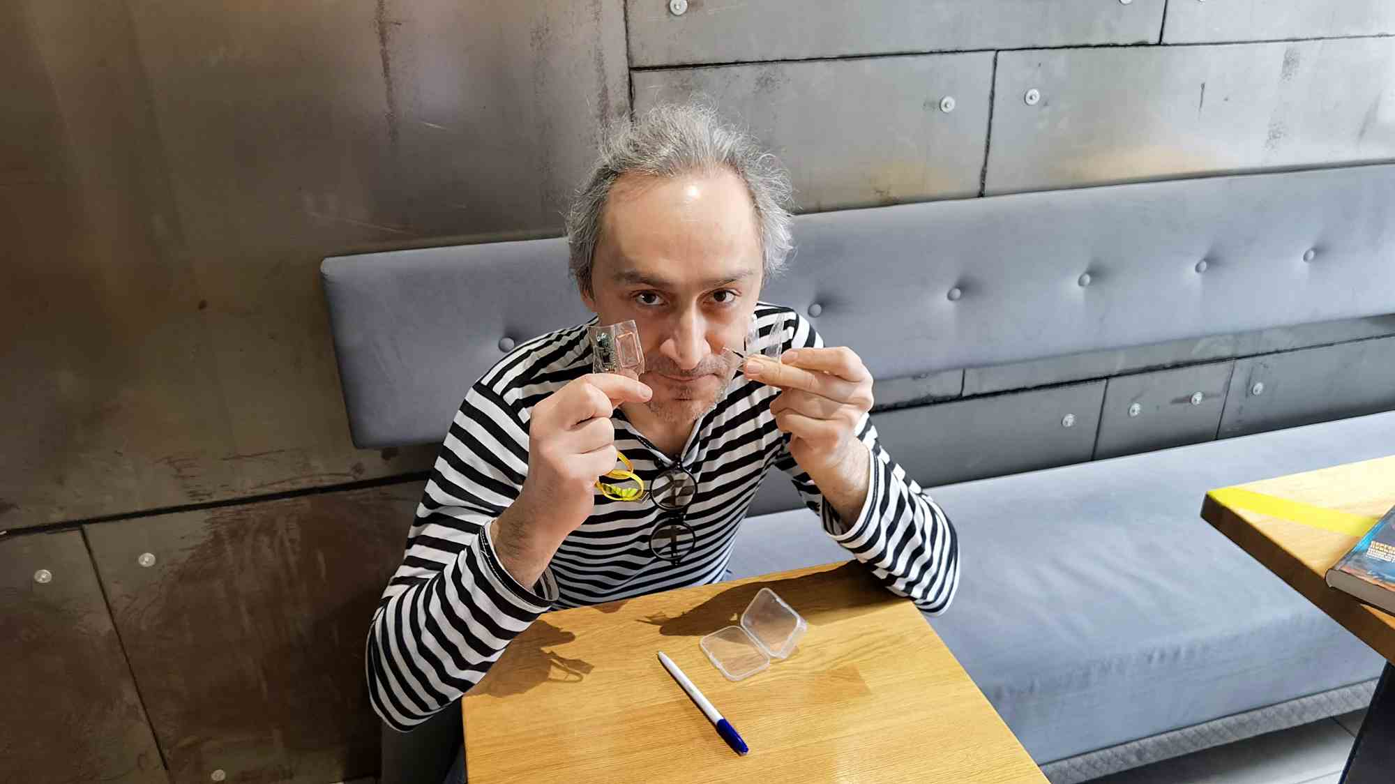 Мужчина в полосатой кофте сидит за столом, держа в руках прозрачные линзы для контактных очков, на столе лежат футляр и ручка.