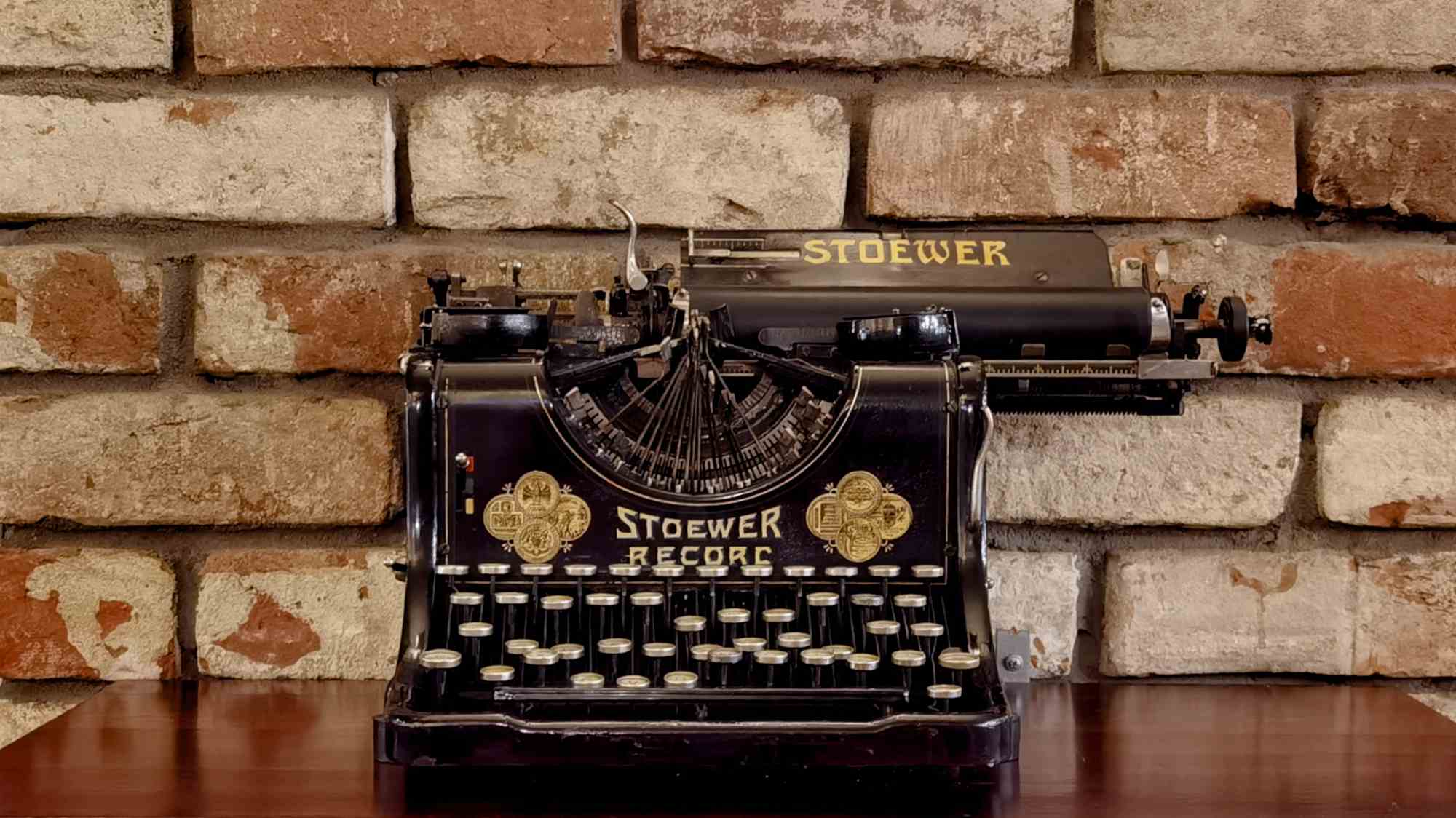 Чёрная пишущая машинка Stoewer Record с золотыми надписями и декоративными элементами стоит на деревянном столе перед кирпичной стеной.