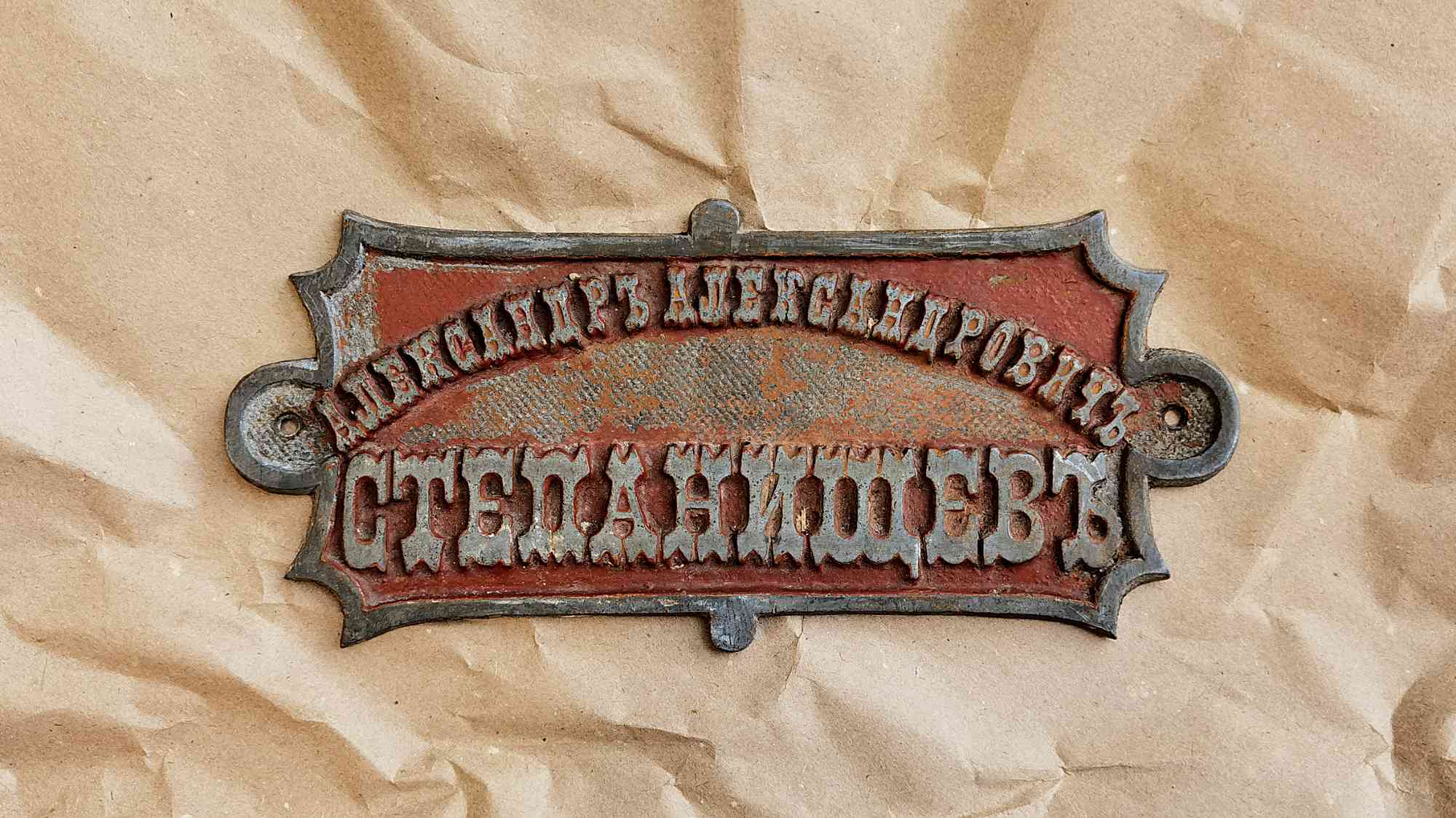 Ржавая металлическая табличка с надписью «АЛЕКСАНДРЪ АЛЕКСАНДРОВИЧЪ СТЕПАНИЩЕВЪ» на фоне мятной бумаги.