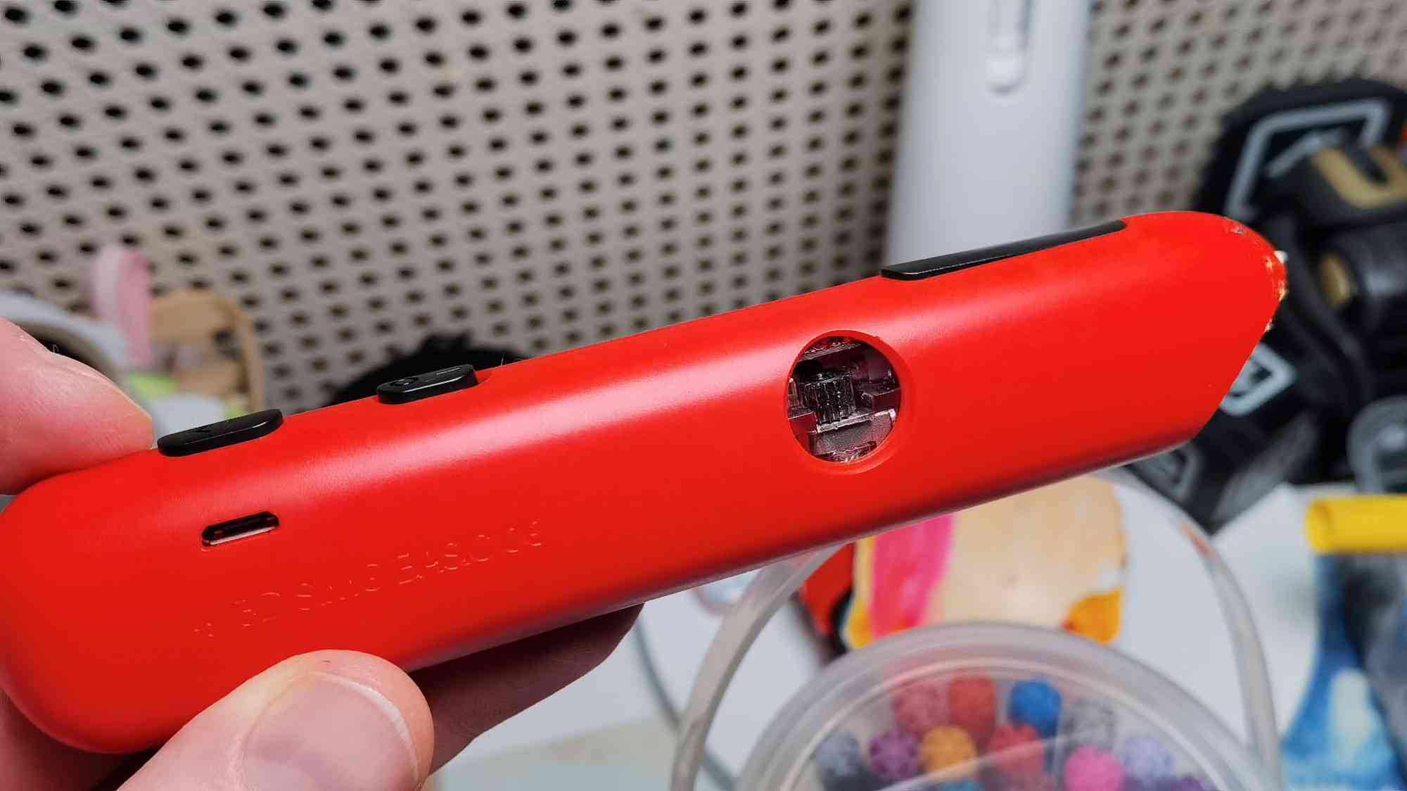 Красный джойстик Nintendo Switch с открытым портом для зарядки, удерживаемый в руке на фоне перфорированной панели и размытых предметов.