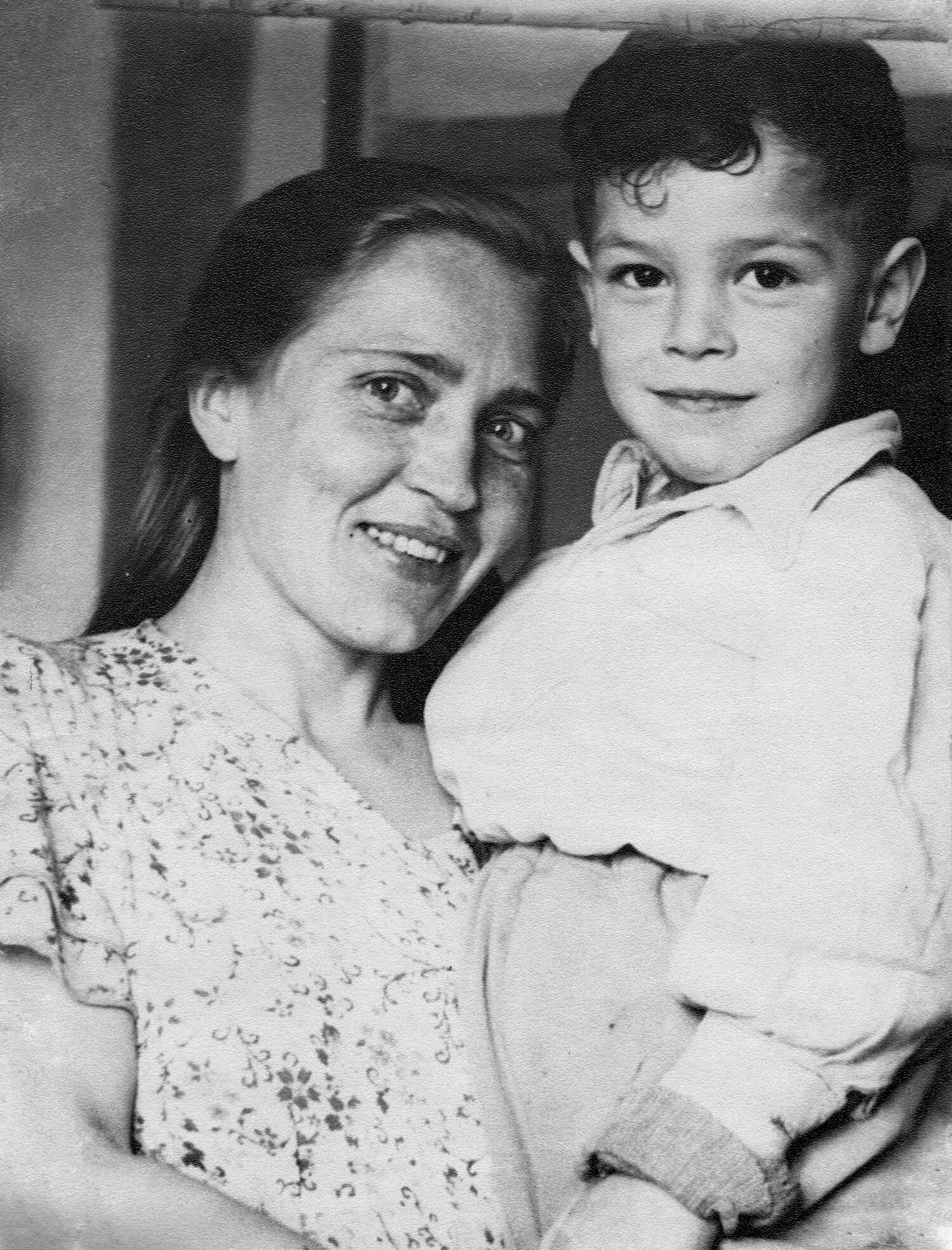 Чёрно-белая фотография: улыбающаяся женщина в цветастом платье держит на руках мальчика в светлой рубашке, оба смотрят в камеру.
