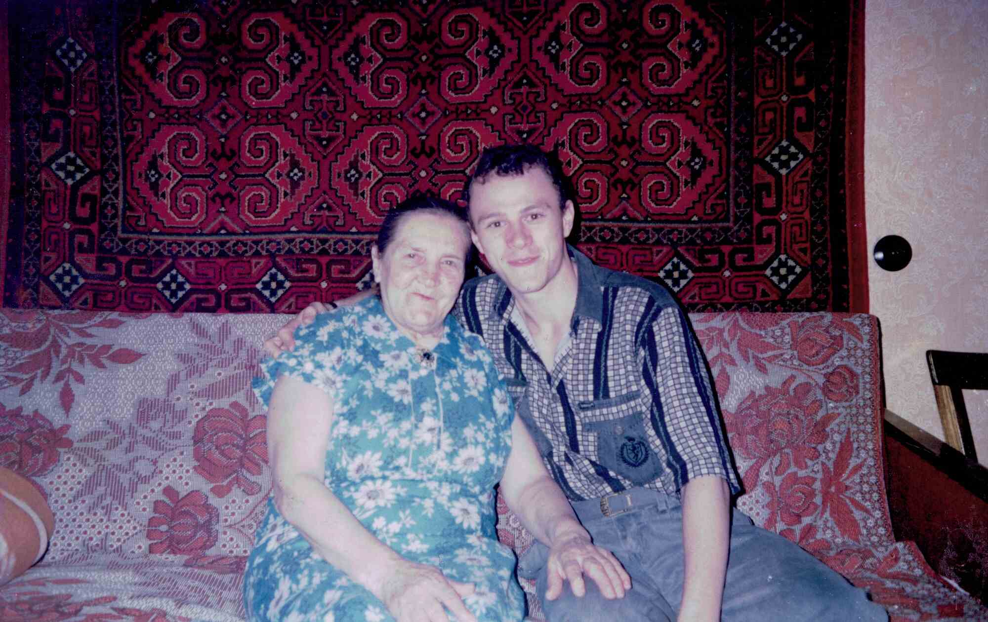 Пожилая женщина в сине-белом цветочном платье и молодой мужчина в клетчатой рубашке сидят рядом на диване с красным узором, на фоне красного ковра с геометрическим орнаментом.