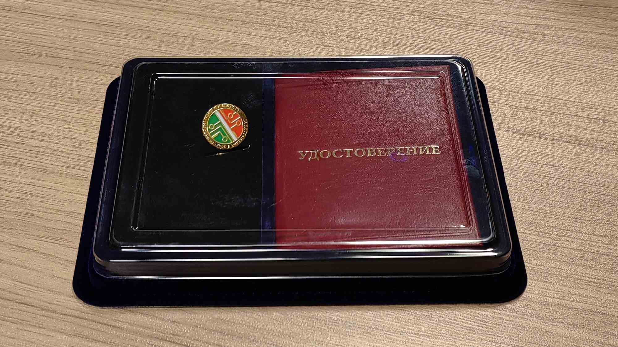 Красное удостоверение с золотым тиснением «УДОСТОВЕРЕНИЕ» и круглая эмблема с зелёно-красным дизайном в чёрном футляре на деревянном столе.