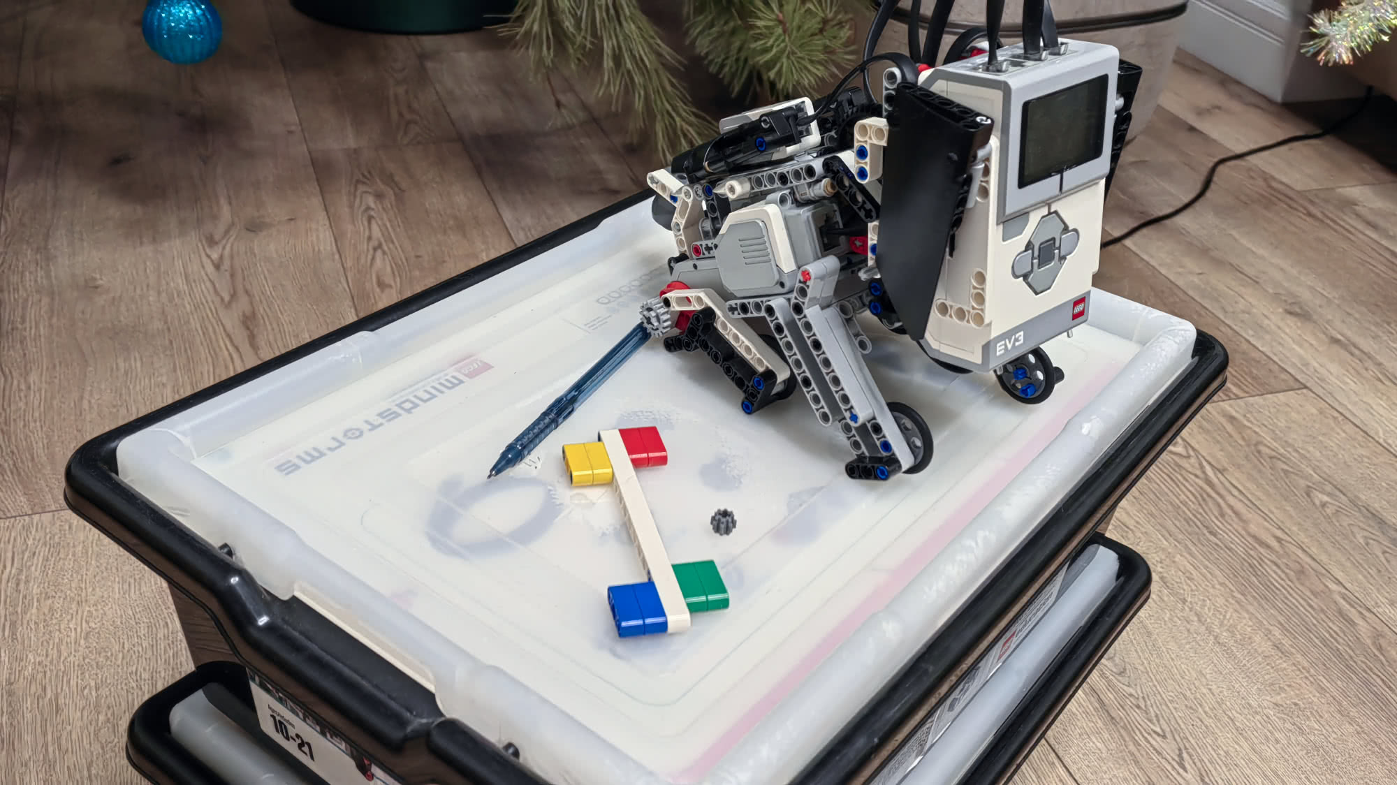Робот LEGO Mindstorms EV3 с ручкой рисует на крышке прозрачного контейнера, рядом лежат цветные кубики и шестерёнка.
