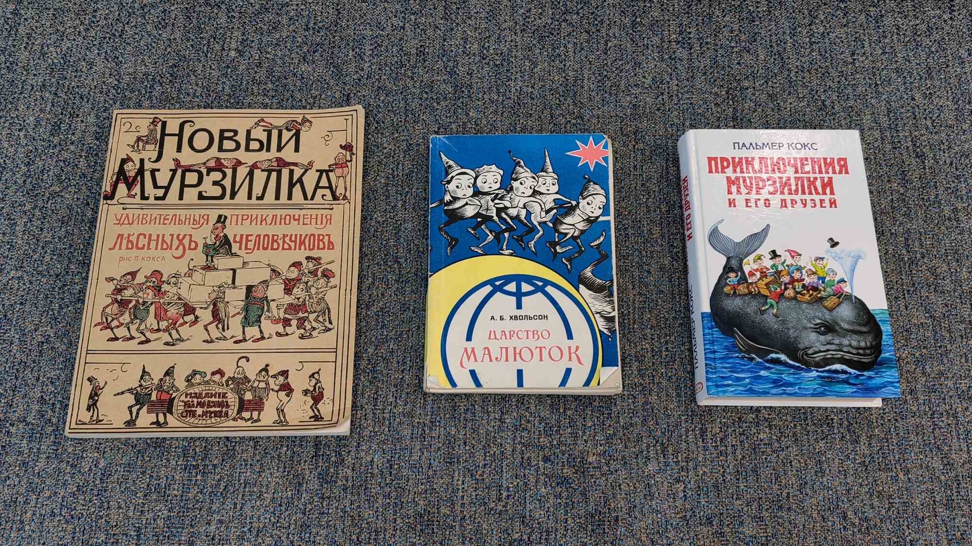 Три детские книги на синем ковре: «Новый Мурзилка» с иллюстрациями П. Кокса, «Царство Малюток» А. Б. Хвольсон и «Приключения Мурзилки и его друзей» Пальмера Кокса.