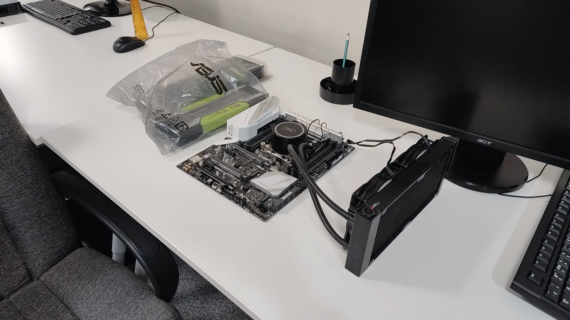 Материнская плата ASUS с установленным жидкостным охлаждением и радиатором на белом столе рядом с монитором Acer и упаковкой компонентов.