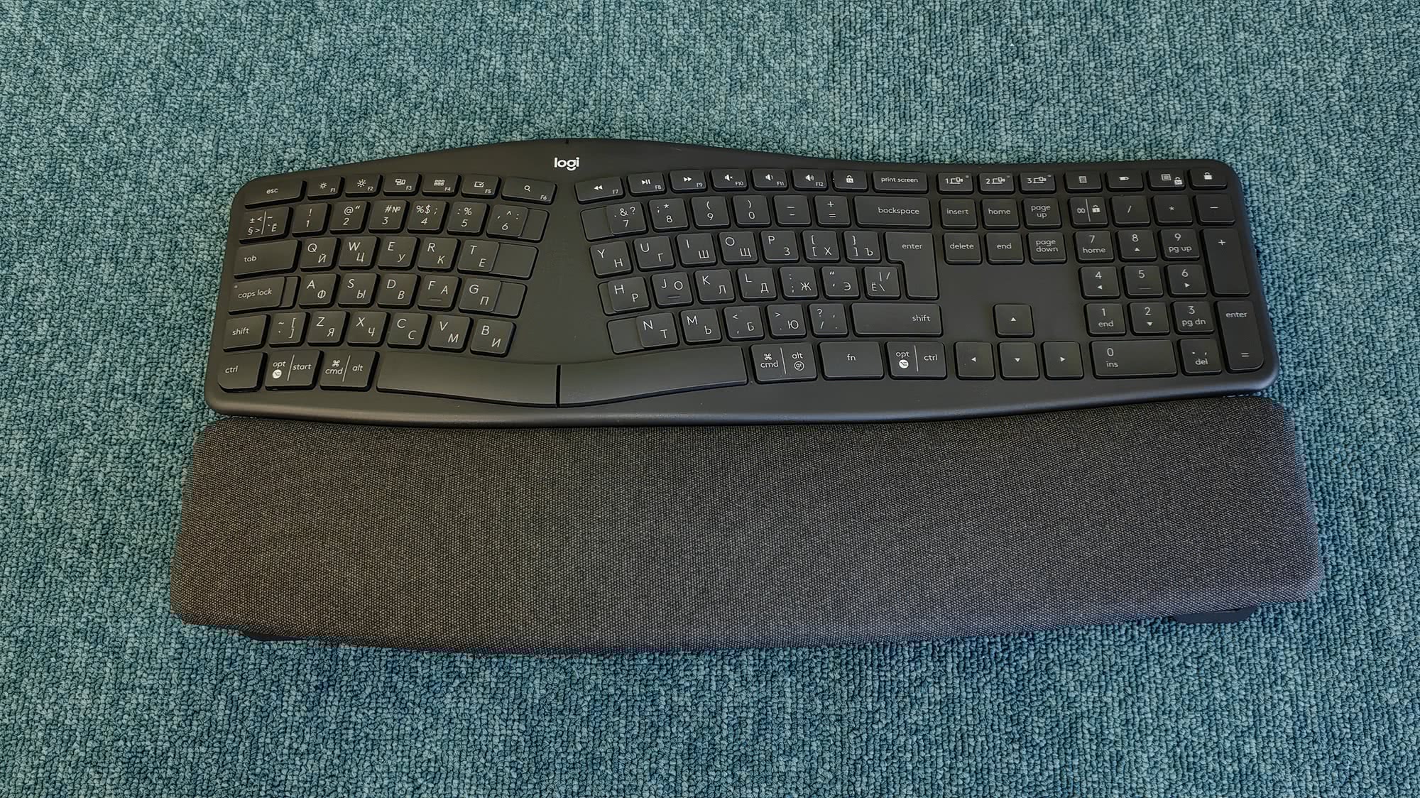 Эргономичная беспроводная клавиатура Logitech с раздельными секциями и подставкой для запястий, расположенная на синем ворсистом ковре.