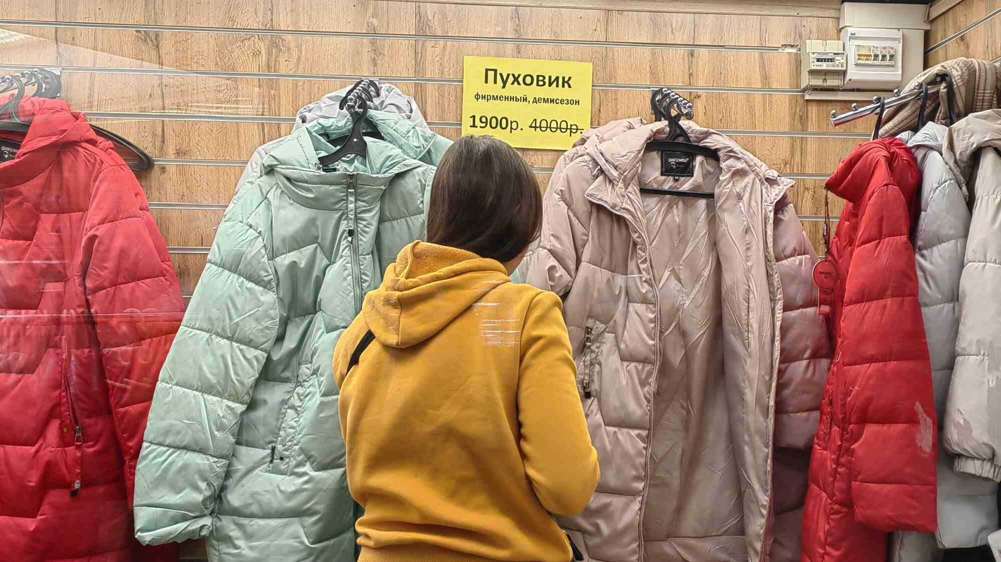 Женщина в жёлтом худи рассматривает пуховики на вешалках; жёлтый ценник: «Пуховик фирменный, демисезон 1900р. 4000р.