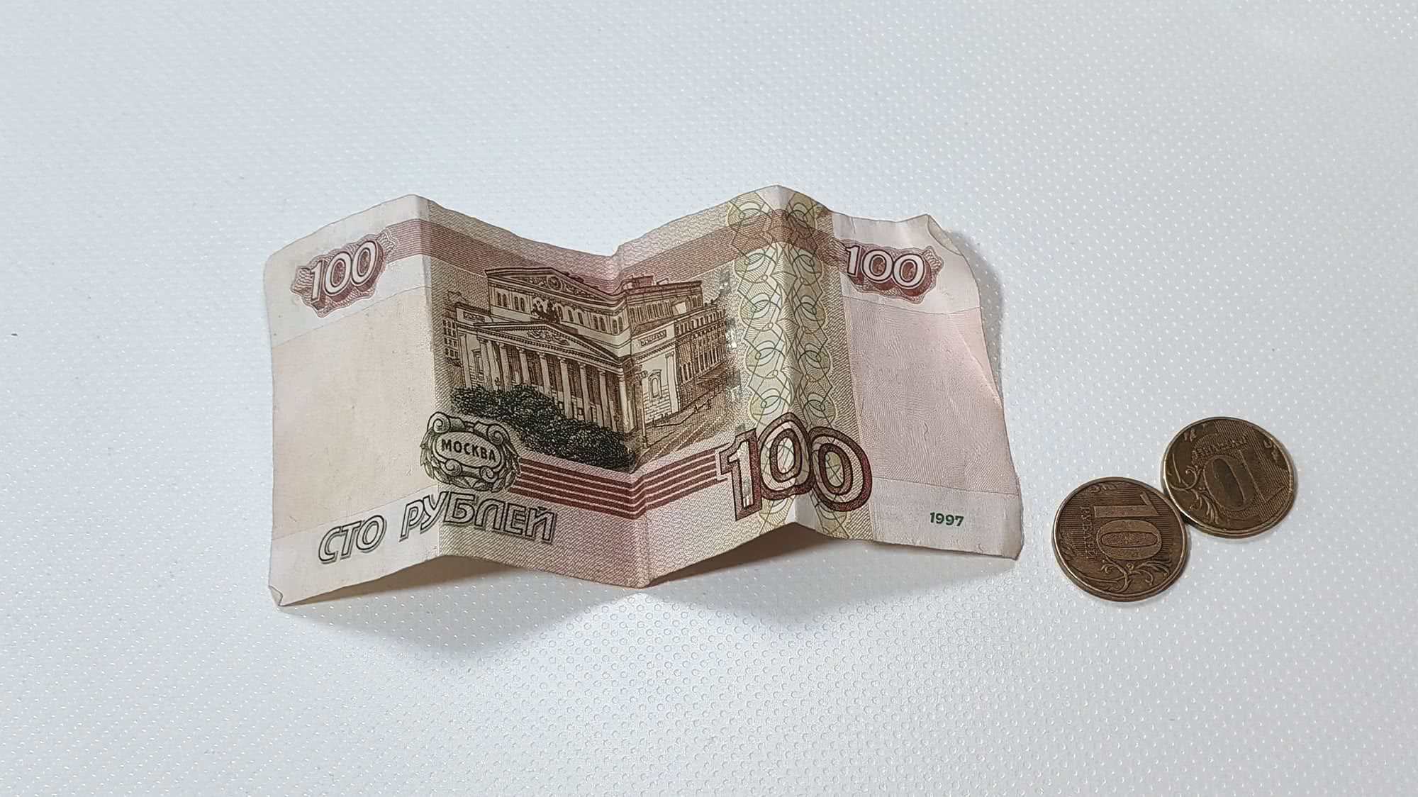 Смятая купюра 100 рублей 1997 года с изображением Большого театра в Москве и две монеты по 10 рублей на белой текстурированной поверхности.