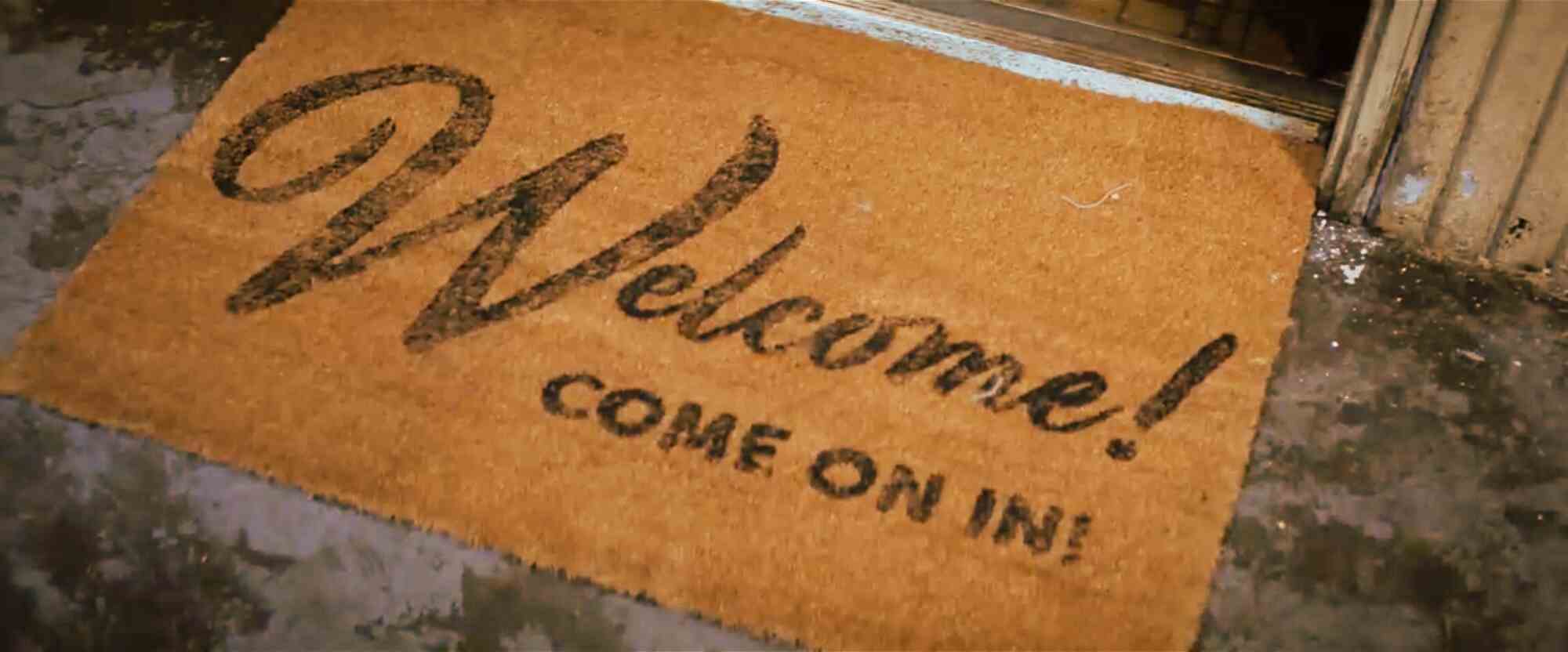 Коврик у входа с надписью «Welcome! COME ON IN!» на фоне бетонного пола и дверного проёма.