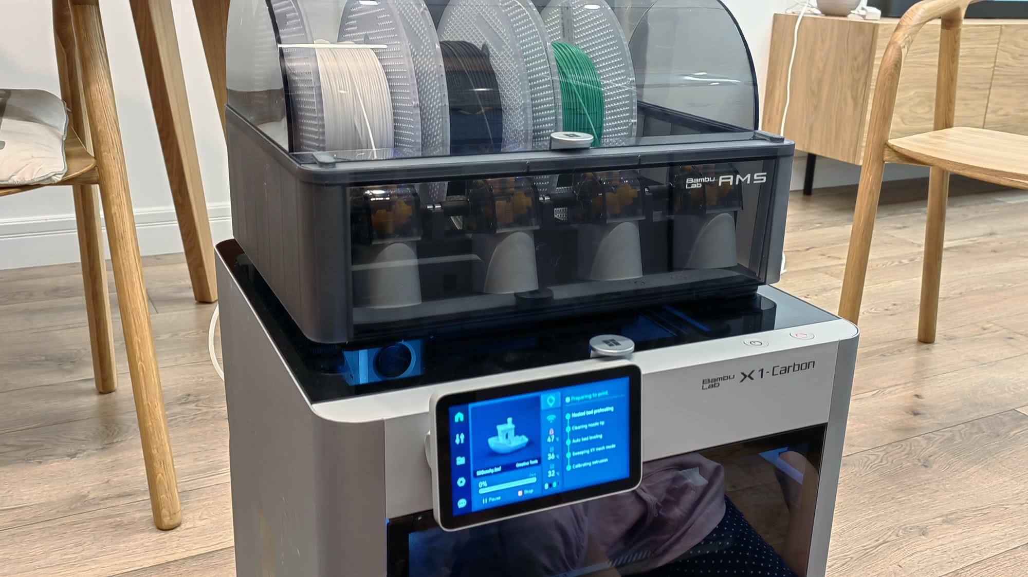 3D-принтер Bambu Lab X1-Carbon с модулем AMS, на экране отображается процесс печати: подготовка стола, очистка сопла, калибровка.