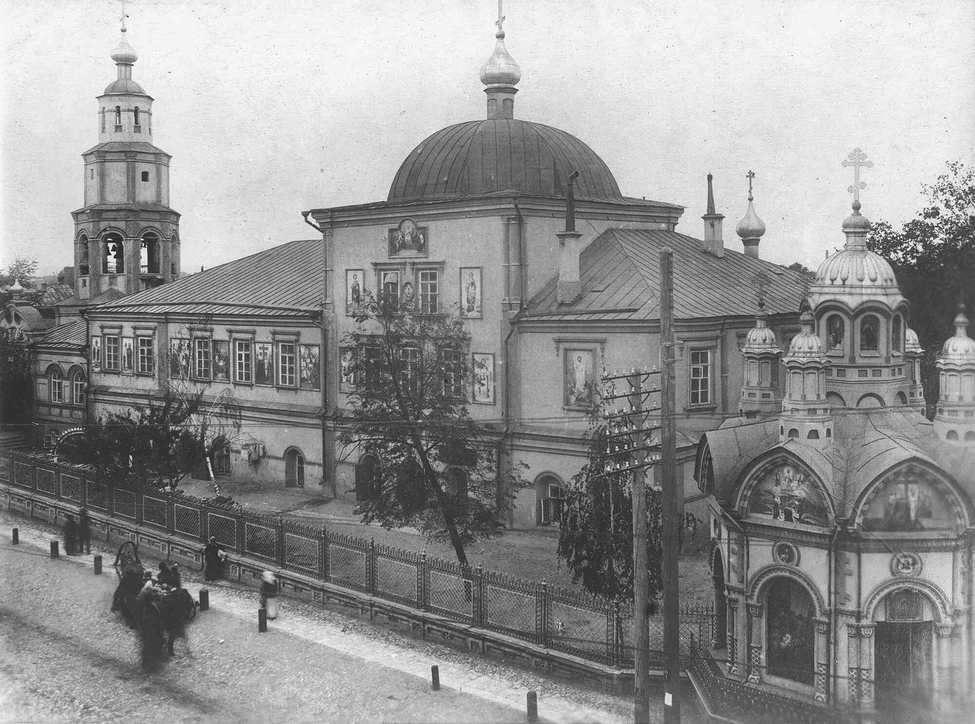 Чёрно-белая фотография старинного русского монастырского комплекса с колокольней, главным храмом под куполом и приделом с луковичными главами; у ограды — конная повозка и пешеходы.