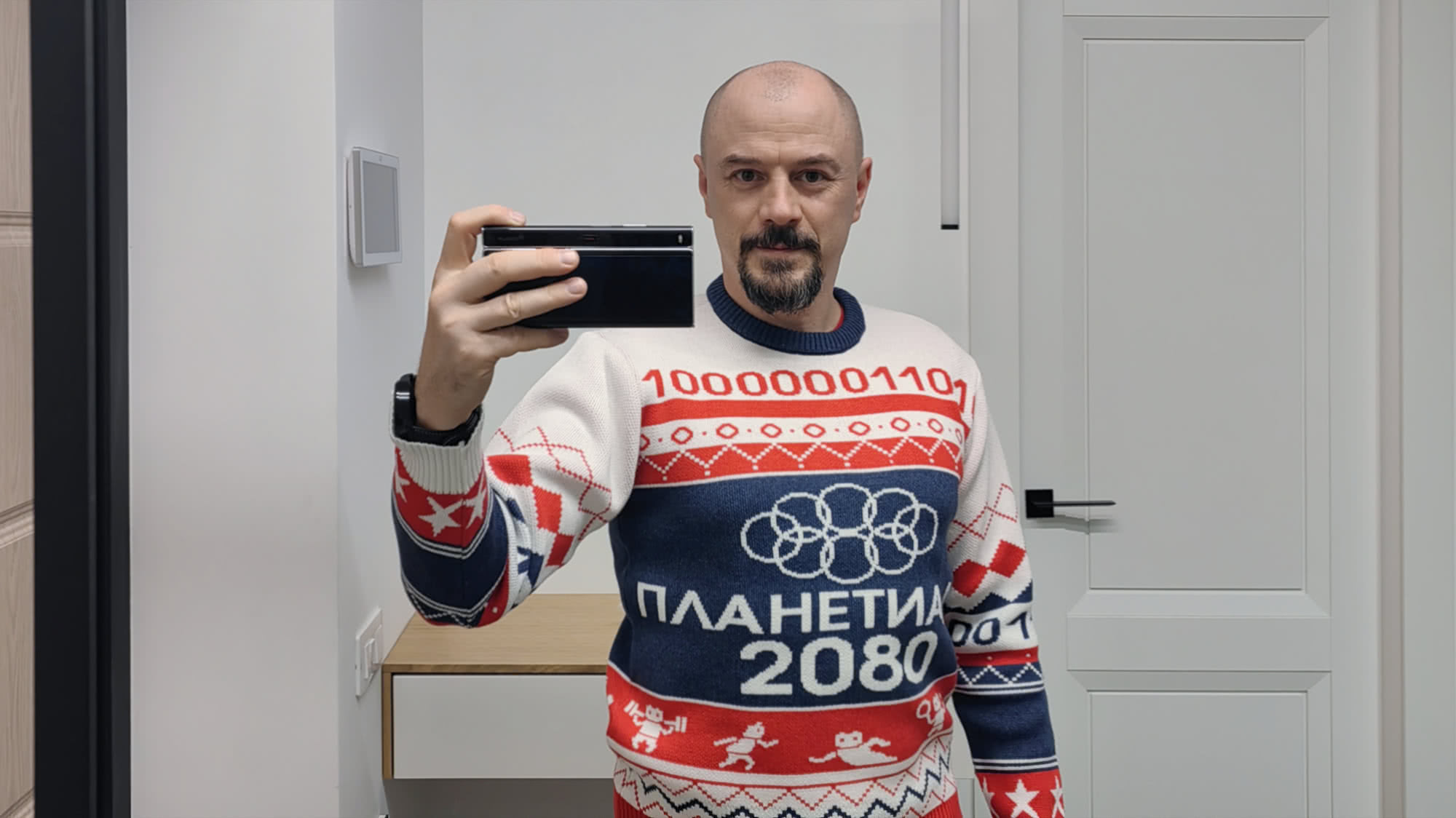 Мужчина в свитере с надписью «ПЛАНЕТИА 2080» и бинарным кодом «10000001101», делает селфи в зеркало, держа смартфон.
