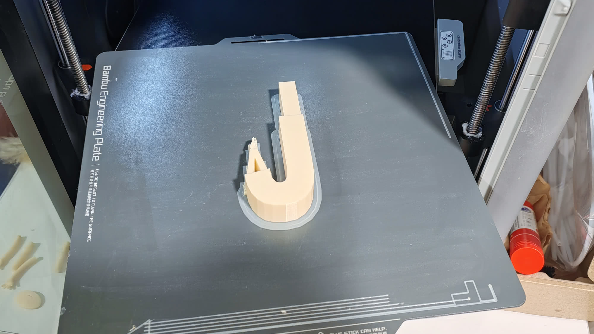 Бежевая 3D-печатная деталь U-образной формы на серой платформе Bambu Engineering Plate с инструкцией по очистке.