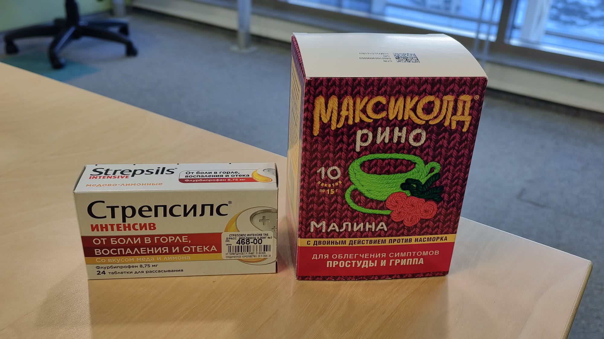 На деревянном столе стоят две упаковки лекарств: белые таблетки «Стрепсилс Интенсив» от боли в горле и красный пакетик «Максиколд Рино» малина от симптомов простуды и гриппа.