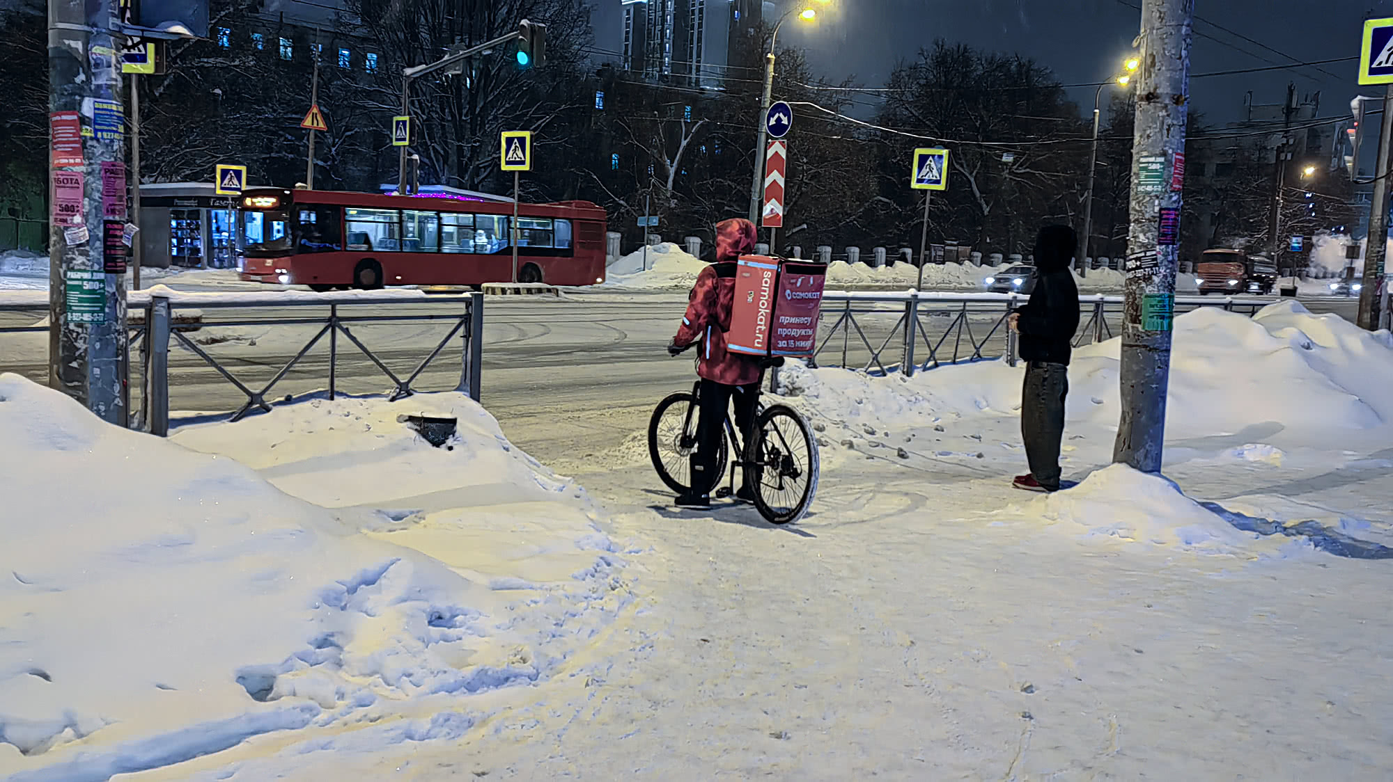 Курьер в красной куртке с рюкзаком Samokat на велосипеде стоит на заснеженной дороге ночью, рядом пешеход; на заднем плане — красный автобус и дорожные знаки.