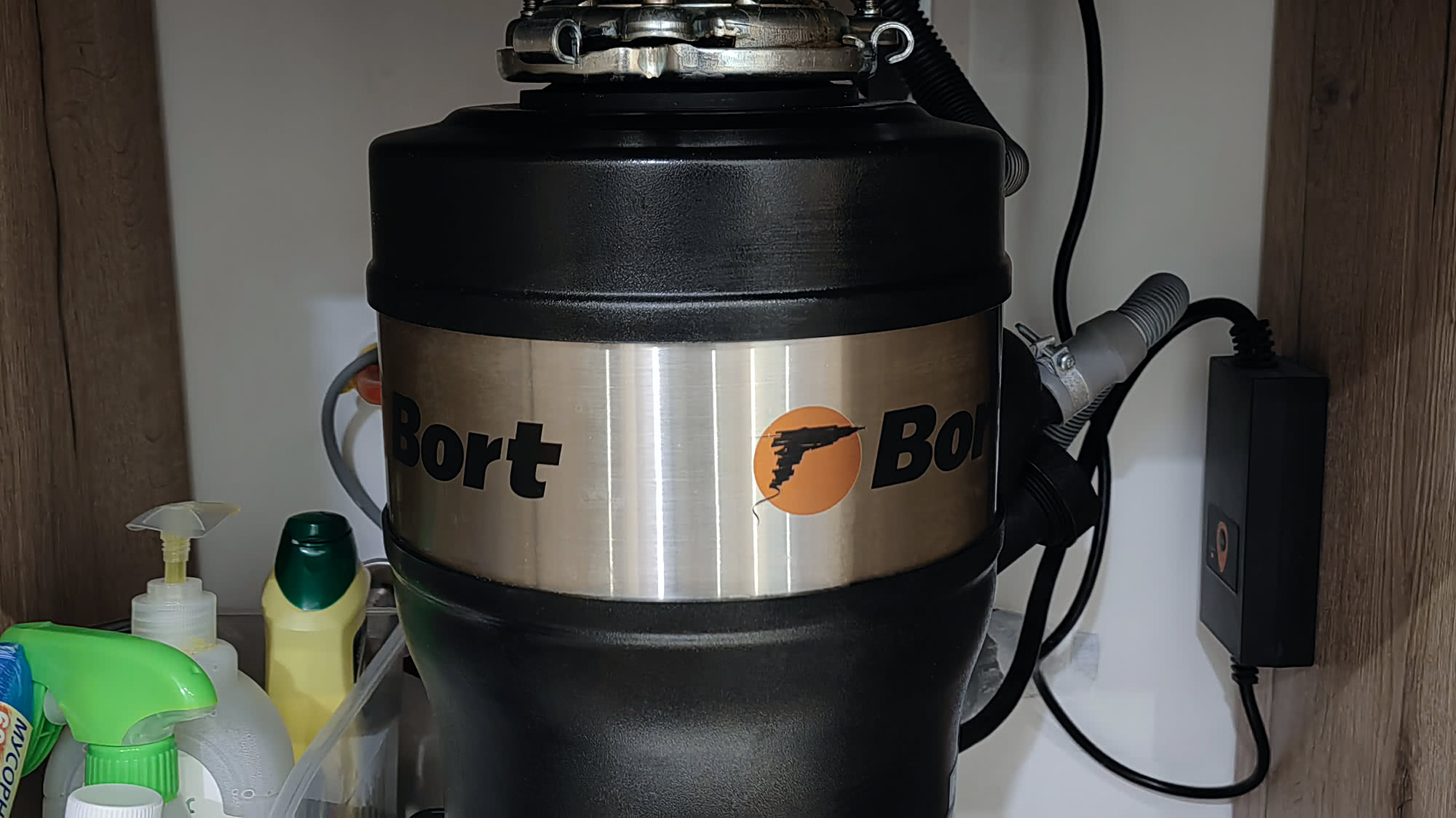 Установленный под раковиной измельчитель отходов Bort с металлическим корпусом, подключённый к электросети и канализации, на фоне бытовой химии.