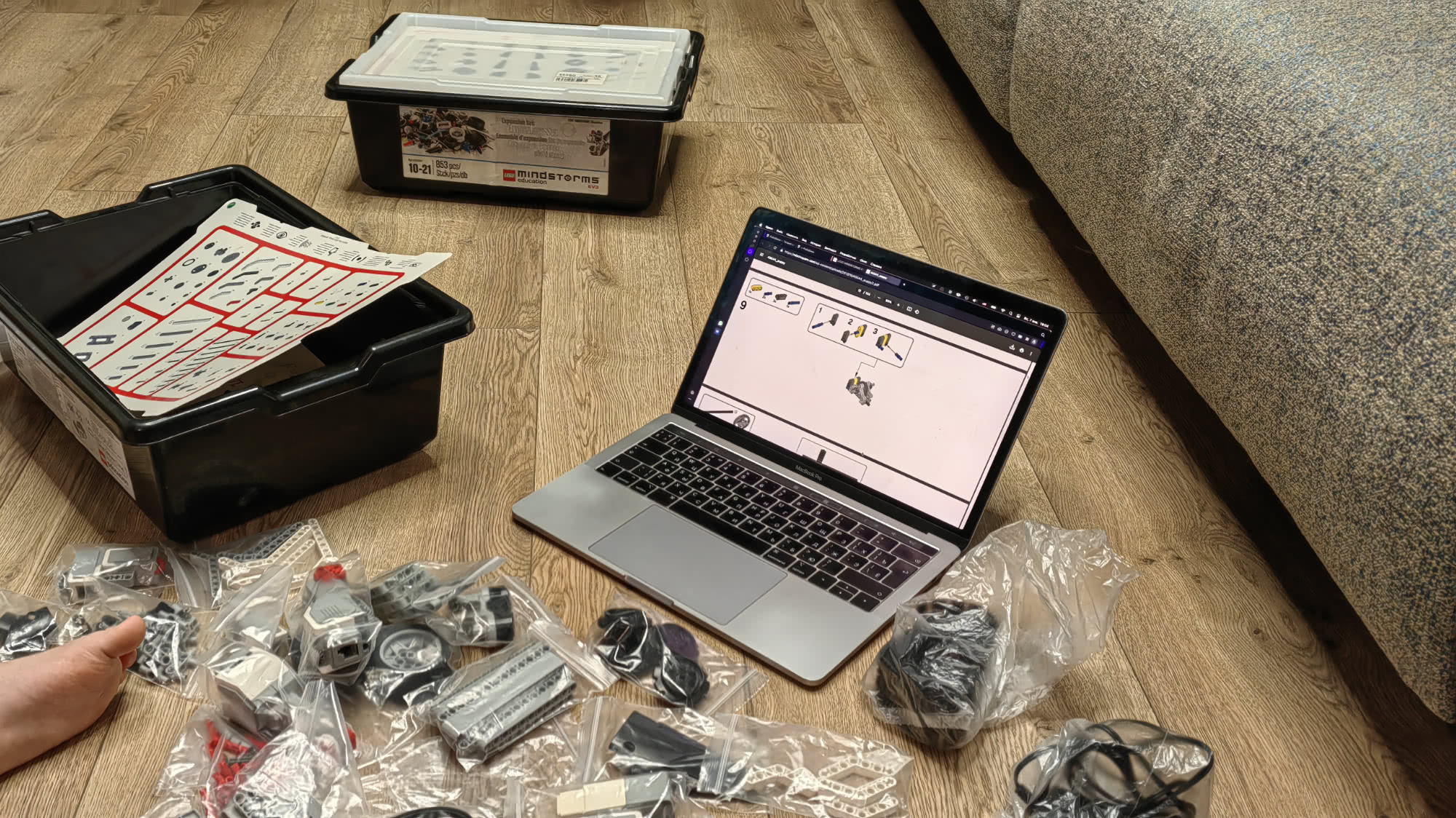 На полу разобраны детали конструктора LEGO Mindstorms Education EV3, рядом открыт ноутбук с инструкцией по сборке и чёрные пластиковые контейнеры для хранения.