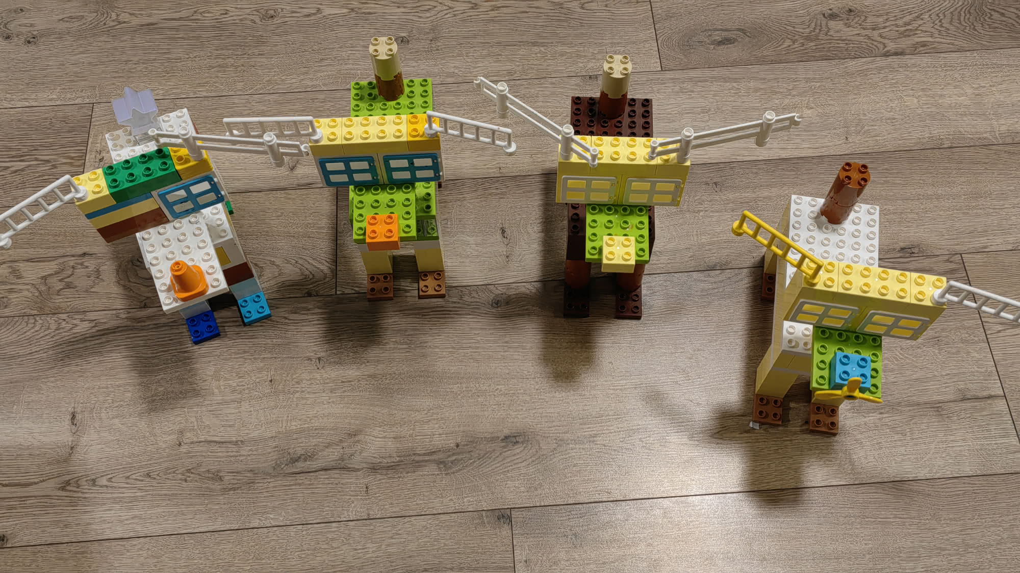 Четыре разноцветные конструкции из кубиков LEGO, напоминающие животных или роботов, стоят на деревянном полу; у некоторых есть лестницы и трубы.