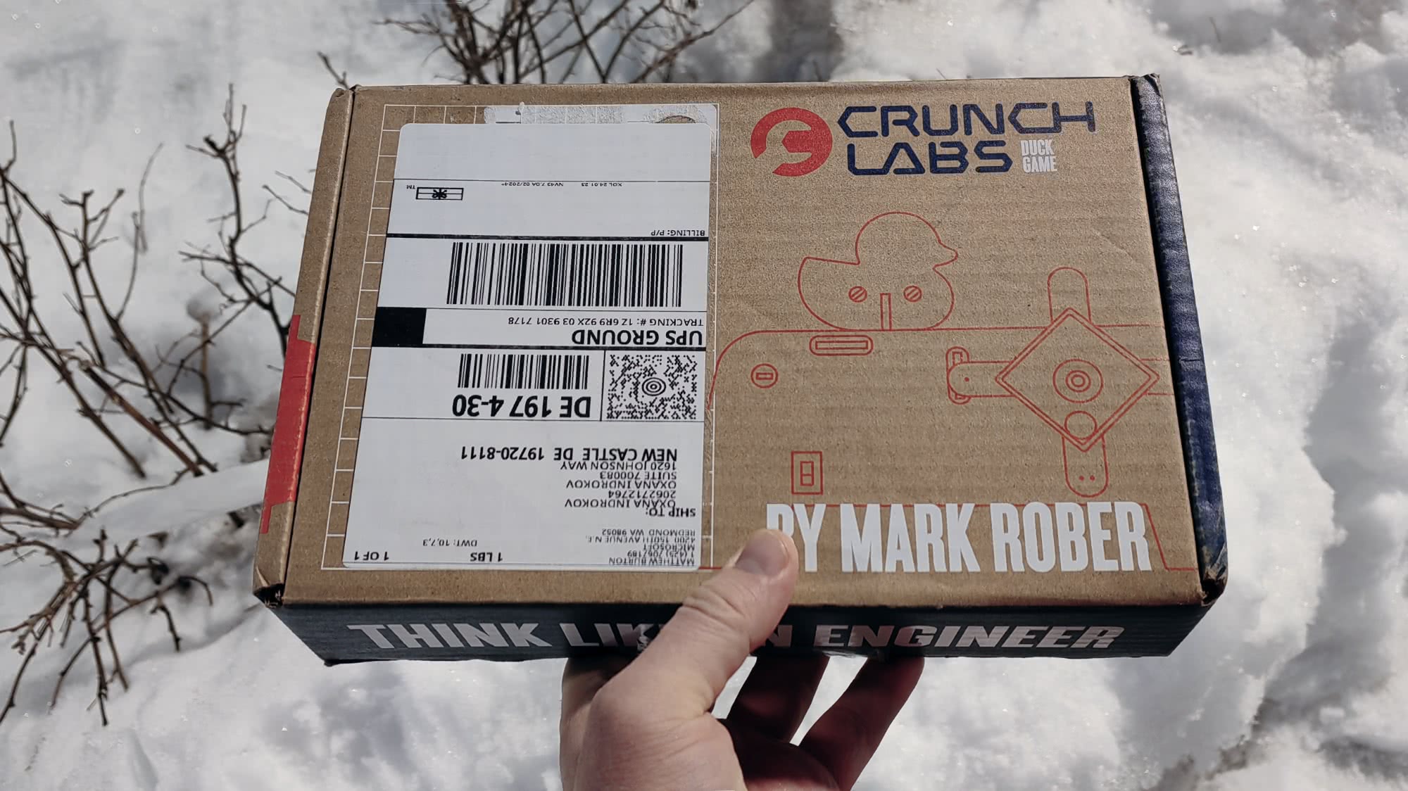 Коробка от Crunch Labs с надписью «BY MARK ROBER» и логотипом Duck Game, держится в руке на фоне снега; видна адресная этикетка UPS Ground.