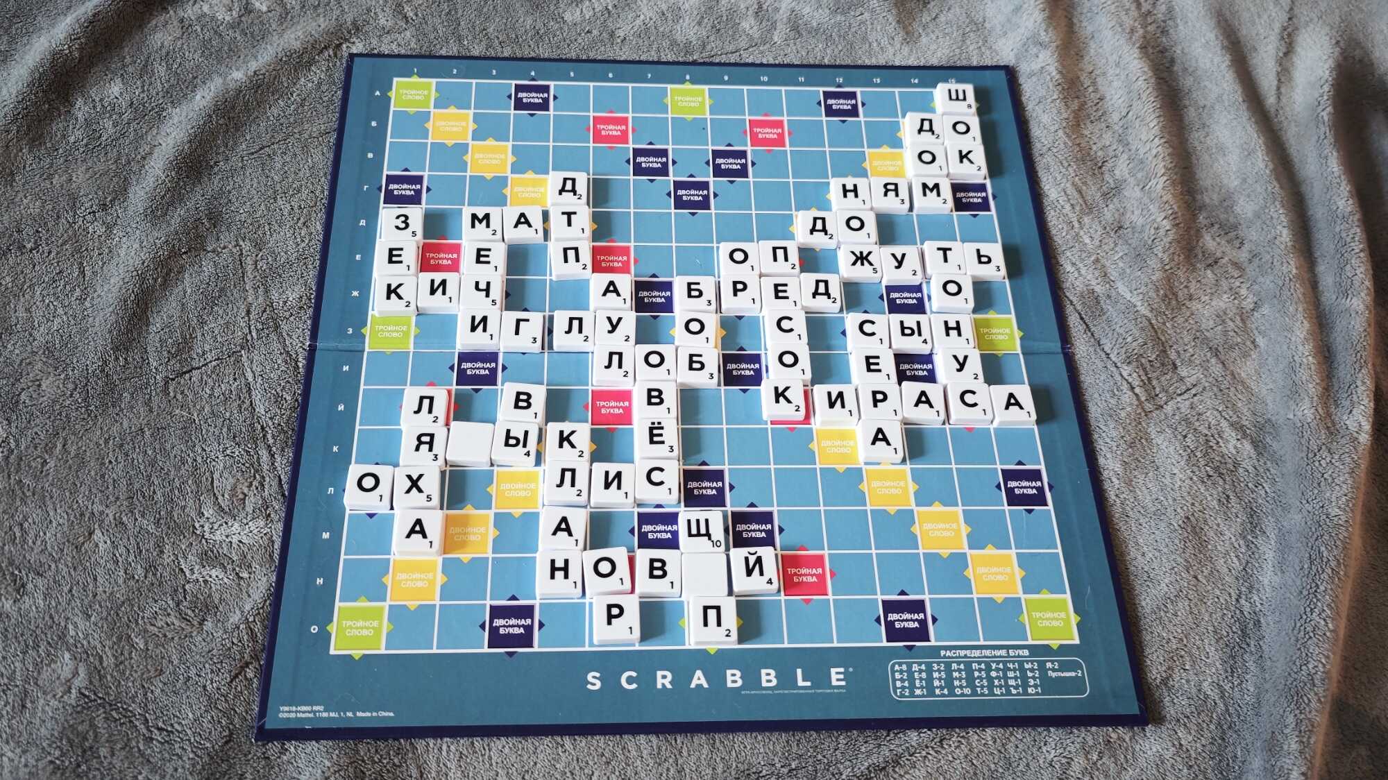 Русская версия игры Scrabble на синем поле с белыми фишками, сложенными в слова: «МАТ», «ДОК», «ЖУТЬ», «КИРКА», «НОВЫЙ» и другие, на сером пледе.