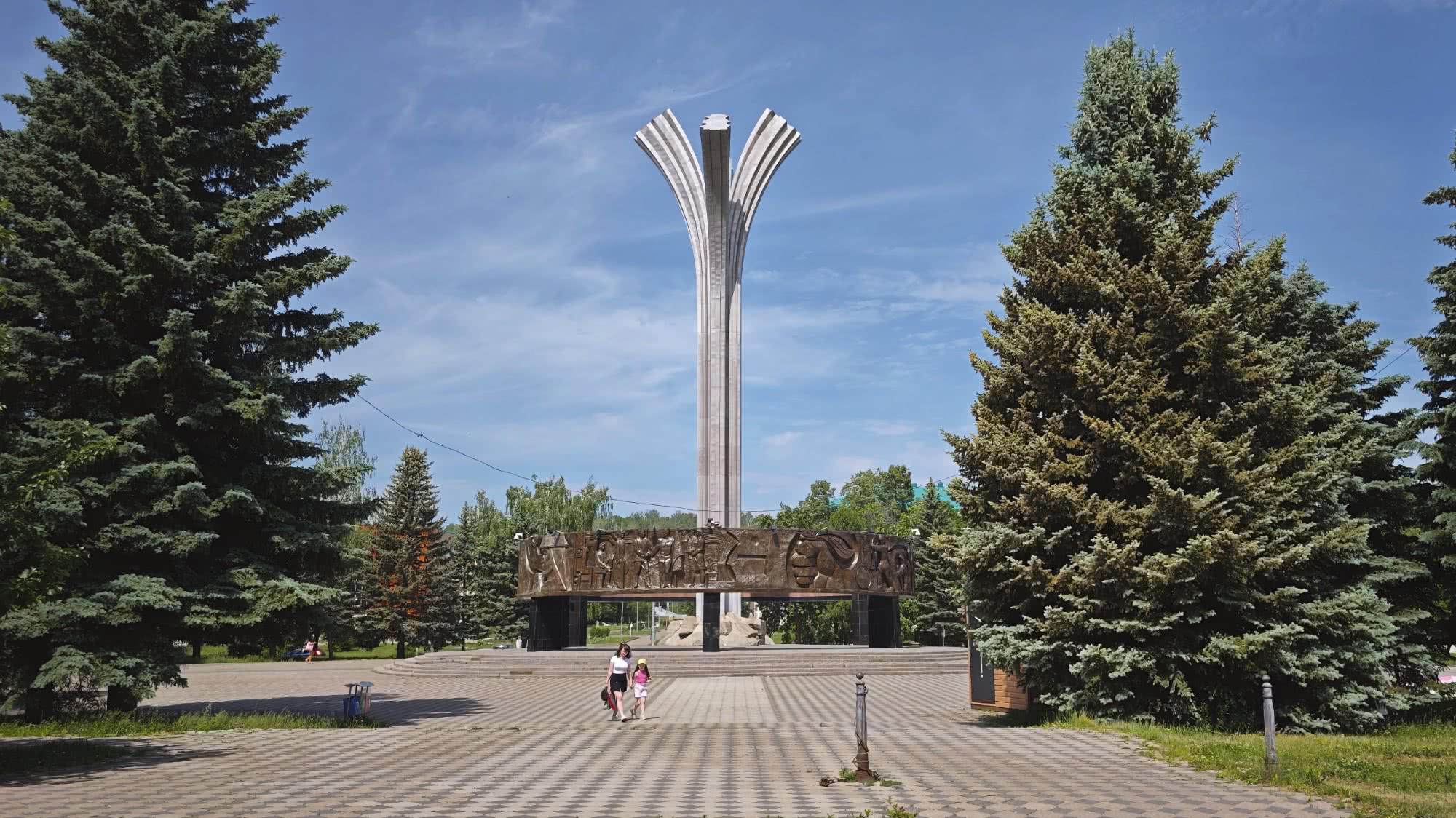 Мемориал «Скорбящая мать» в Новосибирске: высокая стела с тремя лучами, бронзовый барельеф у основания, две женщины идут по площади среди елей под голубым небом.