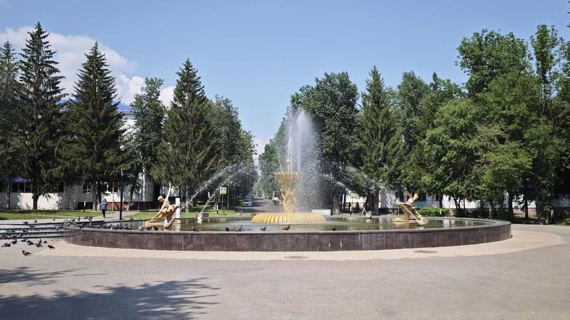 Большой круглый фонтан с золотыми скульптурами и бьющими струями воды, окружённый голубями, деревьями и зданиями на фоне ясного неба.