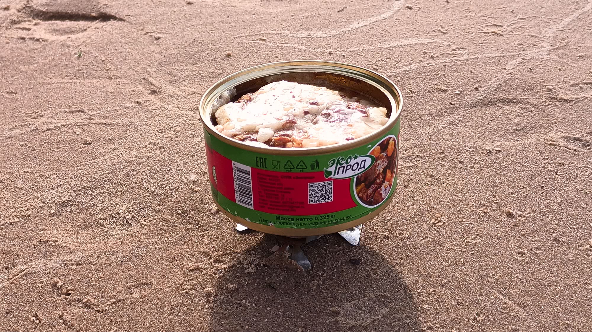Открытая консервная банка с тушёным мясом и жиром, стоящая на песке на подставке; на этикетке видны надписи «ЭкоПрод», «Масса нетто 0,325 кг» и QR-код.