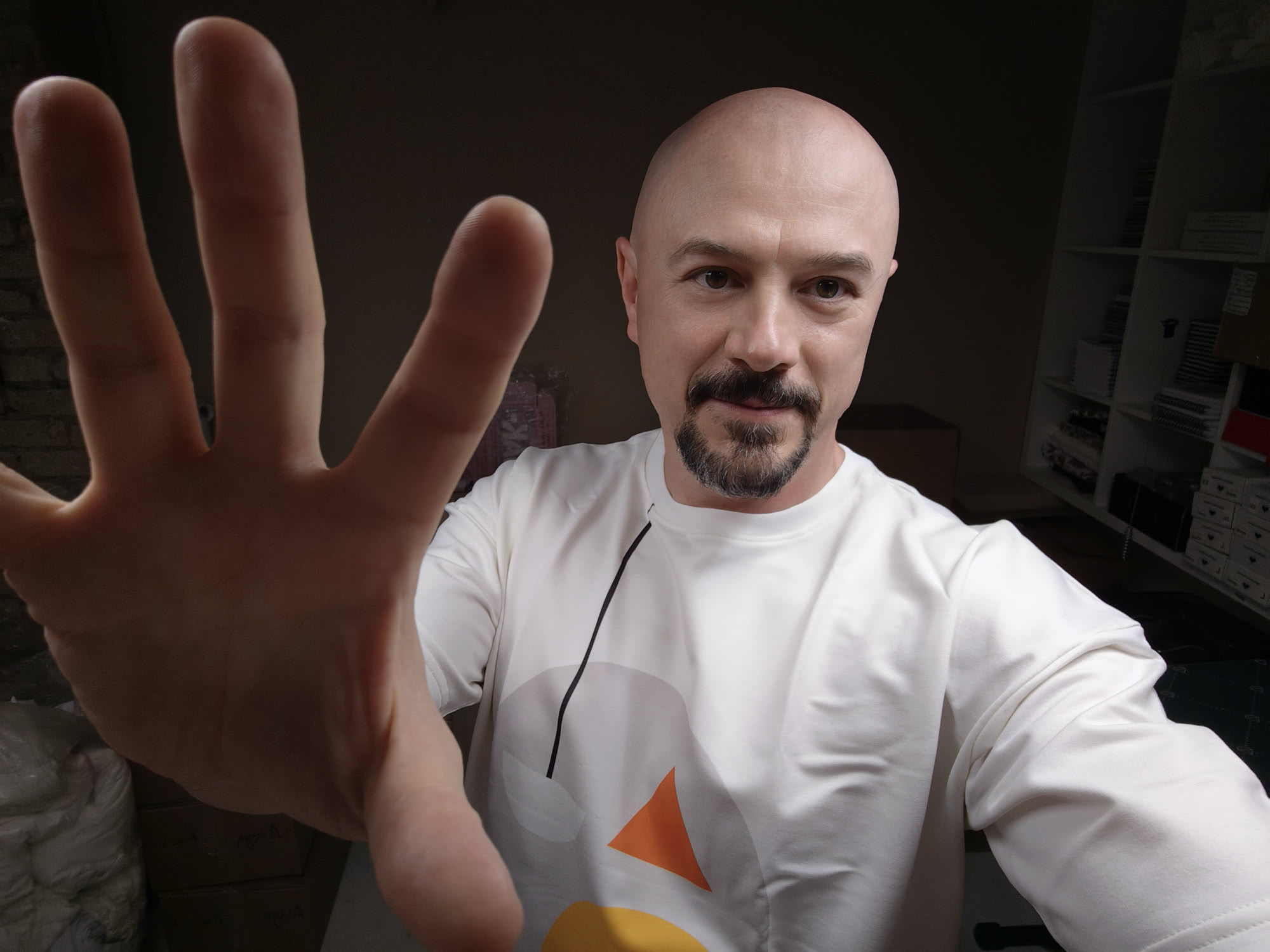 Бородатый лысый мужчина в белой футболке с геометрическим принтом делает селфи, протягивая ладонь к камере; на заднем плане видны стеллажи с коробками.