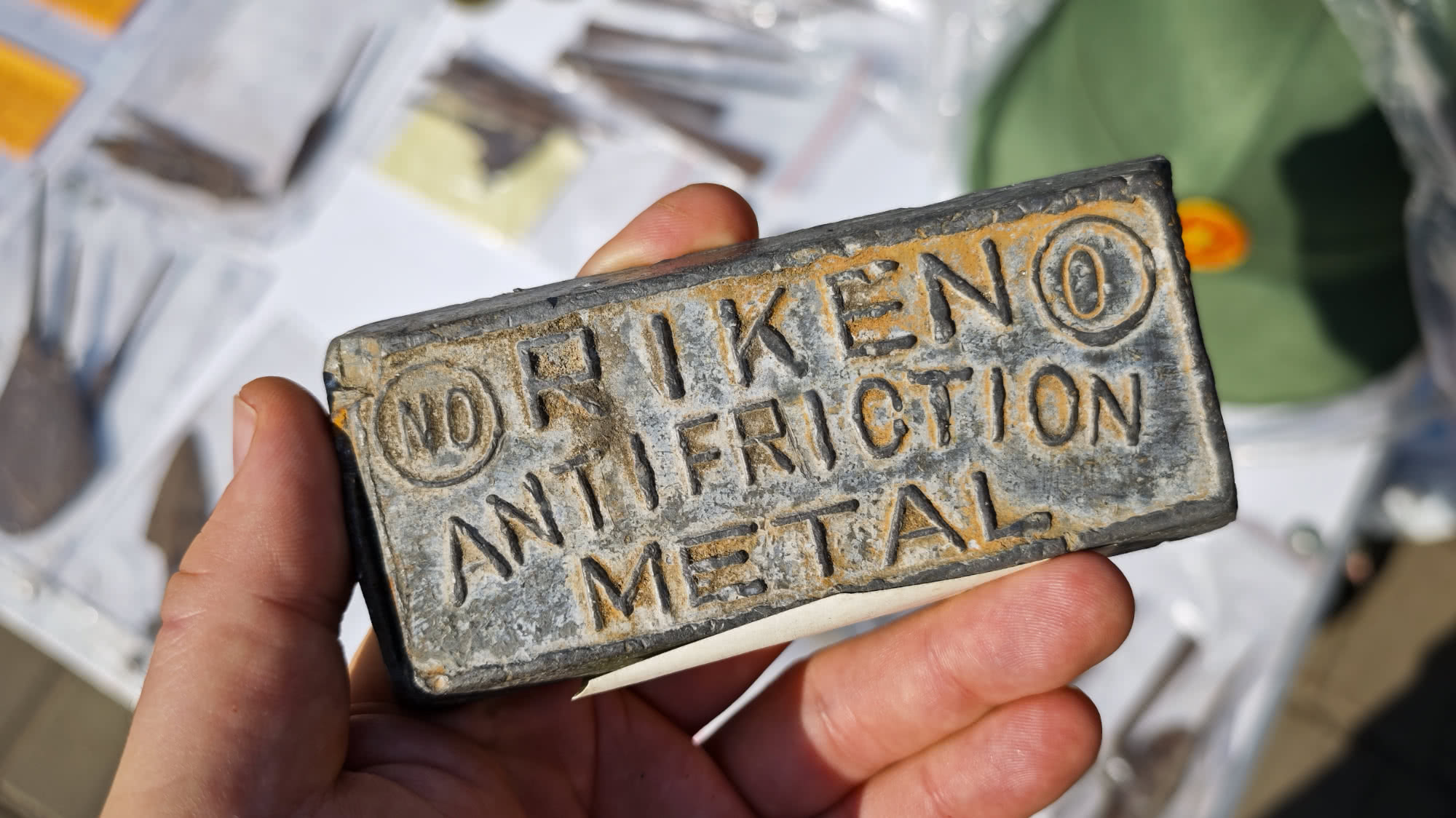 Рука держит прямоугольный металлический брусок с надписью «NORIKENO ANTI FRICTION METAL» и знаком «©», на фоне размытых деталей и упаковки.