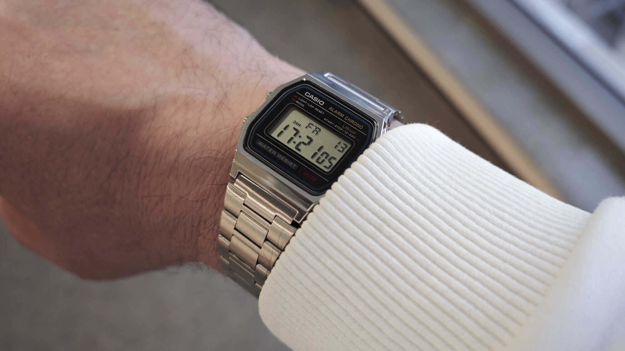 Серебристые цифровые наручные часы Casio с металлическим браслетом на запястье в белом рукаве; на дисплее время 17:21, дата 13.05, день недели FR.