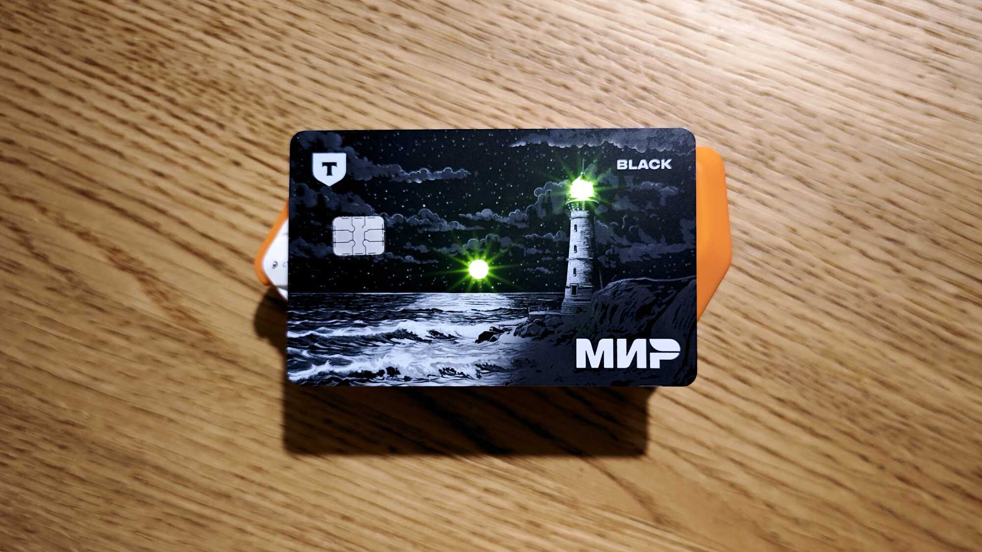 Банковская карта «Мир Black» с изображением маяка на фоне ночного моря и звёздного неба, логотип Т-Банка и чип слева.