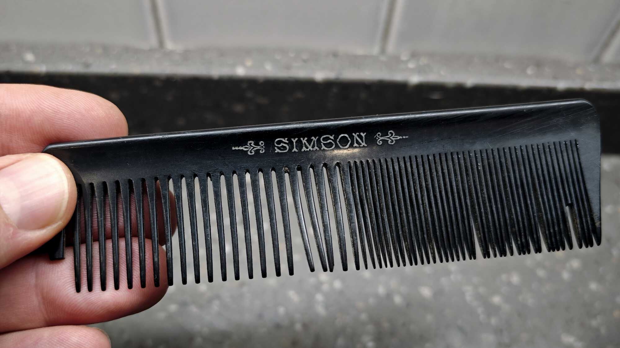 Чёрная расчёска с двойными зубьями, в руке, с надписью «SIMSON» и орнаментами на верхней части.