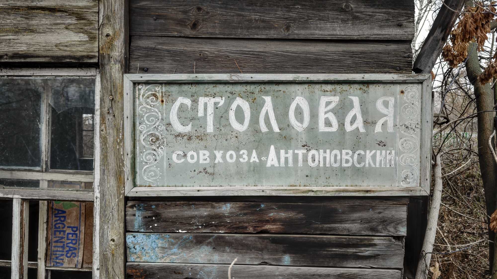 Потёртый металлический указатель с белой надписью «СТОЛОВАЯ» и «совхоза «Антоновский», прикреплённый к деревянной стене старого здания.