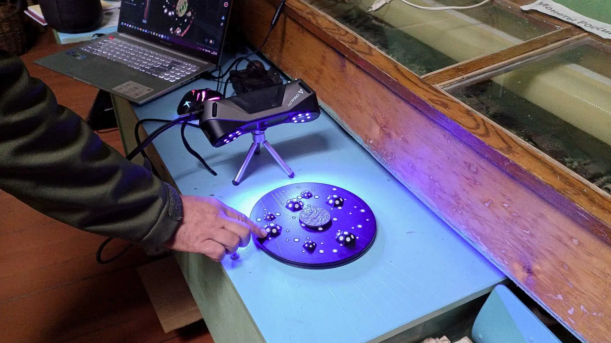 Рука настраивает круглый объект с монетой и точками под фиолетовым светом 3D-сканера Creality на столе рядом с ноутбуком и стеклянным шкафом.