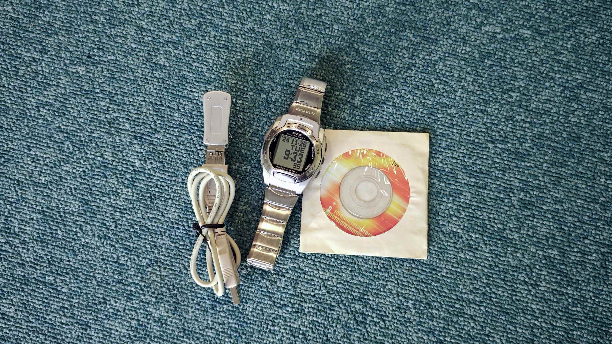 Серебристые цифровые часы Casio Wrist Camera с USB-кабелем и CD-диском в бумажном конверте на синей ковровой поверхности.