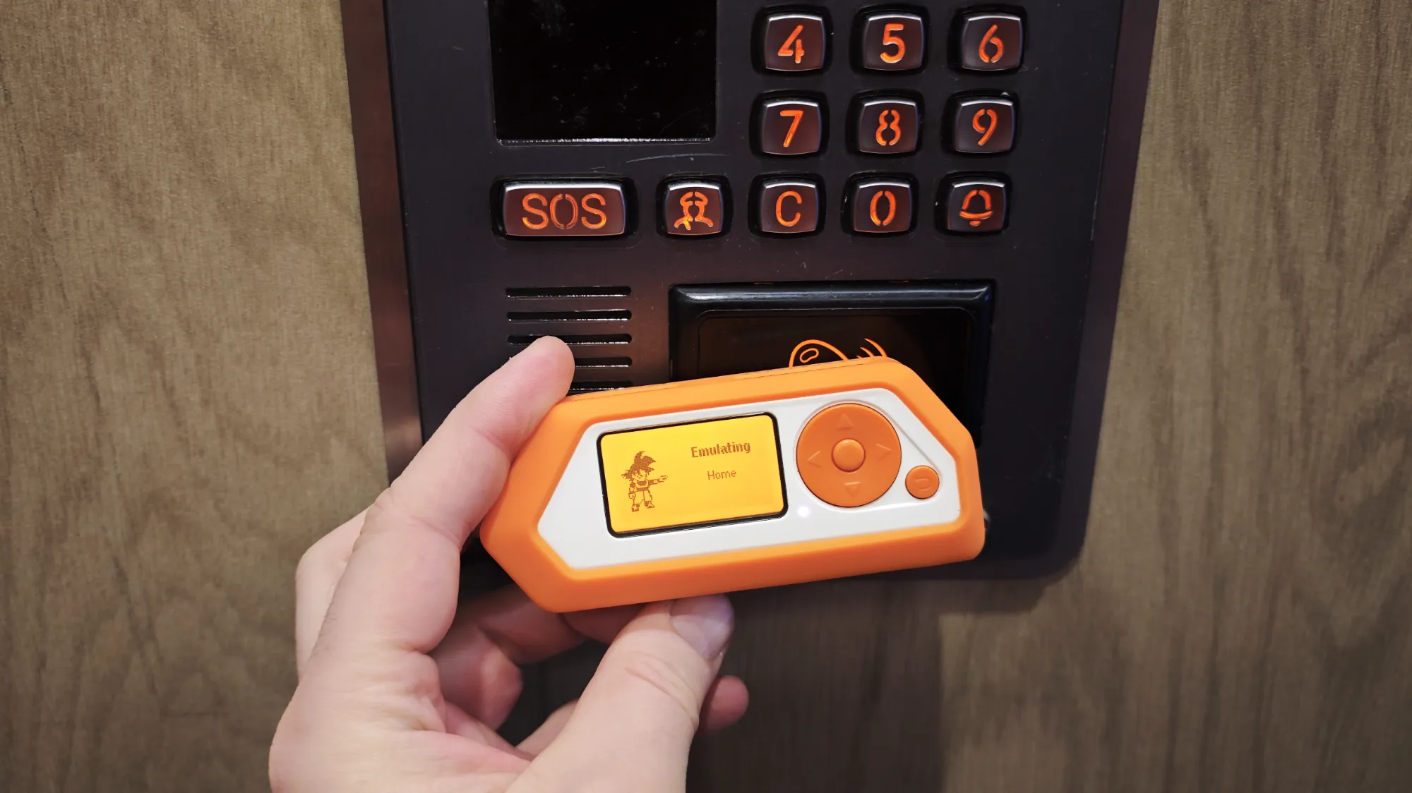 Рука держит оранжево-белый клонировщик ключей с экраном «Emulating Home» у панели домофона с кнопками 0–9, SOS и C.